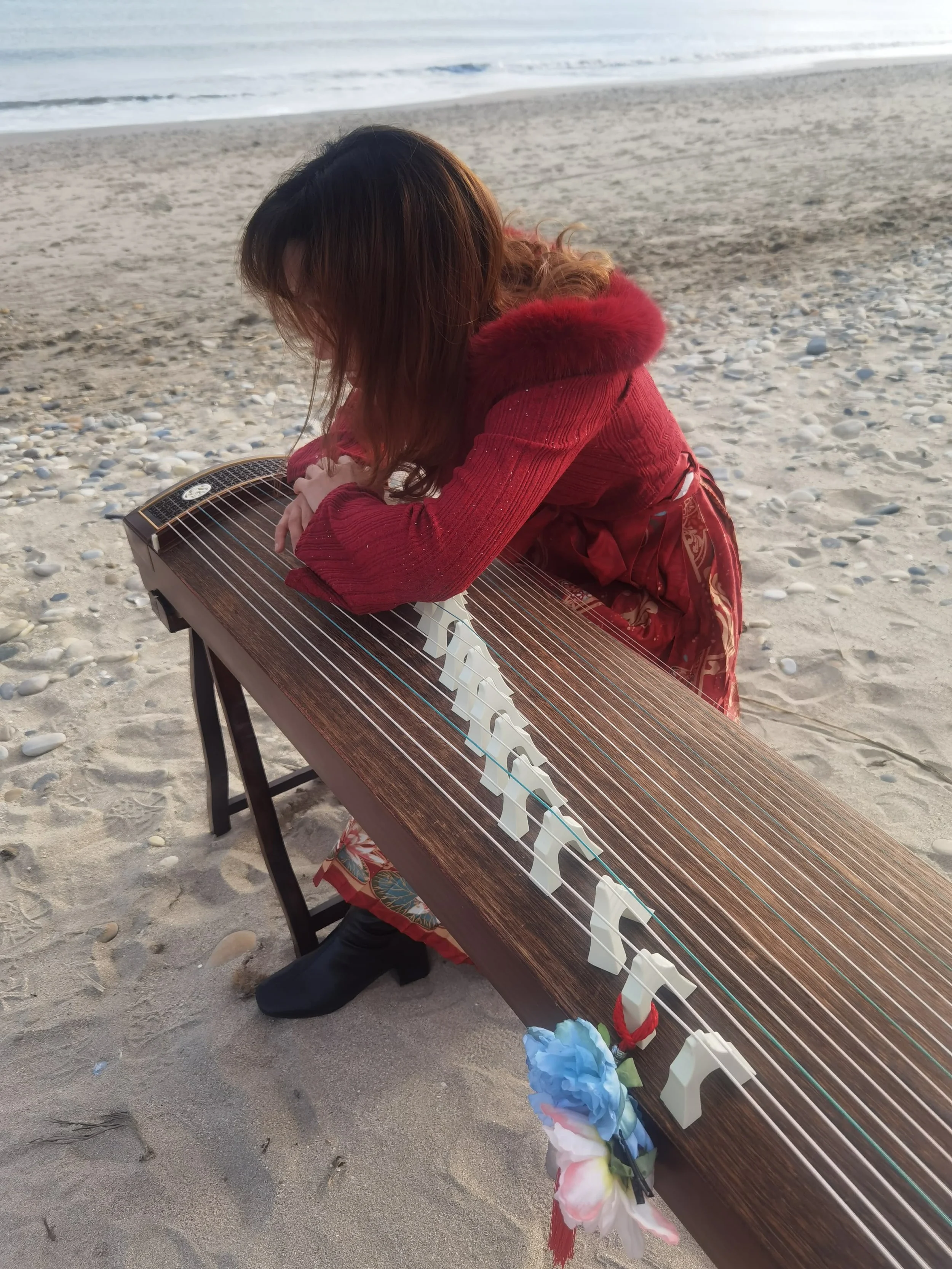 Une femme aux cheveux longs bruns, portant une veste rouge avec une capuche en fourrure rouge, joue de la harpe en position agenouillée sur la plage, face à la mer.