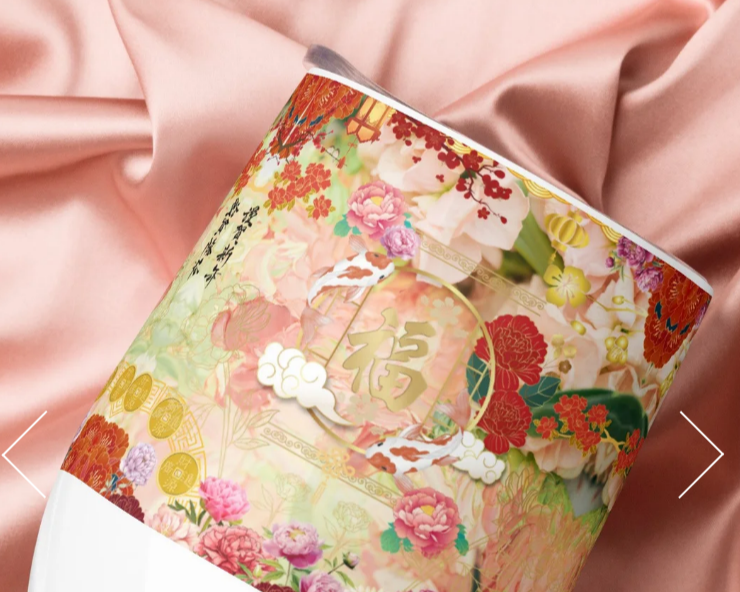 Un verre blanc avec une illustration artistique comprenant des poissons, des fleurs et des motifs dorés, posé sur un tissu rose en satin
