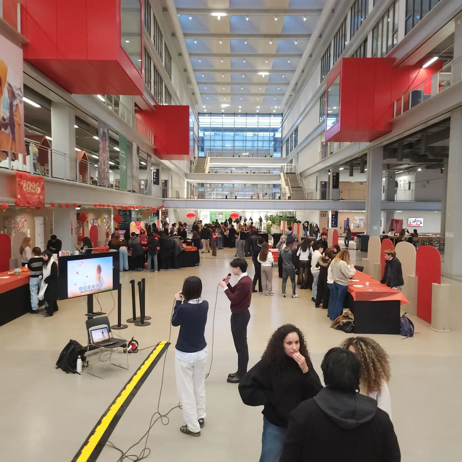 Intérieur d'un grand centre commercial ou espace d'événement avec de nombreuses personnes, décorations rouges, écran et tables d'activité.