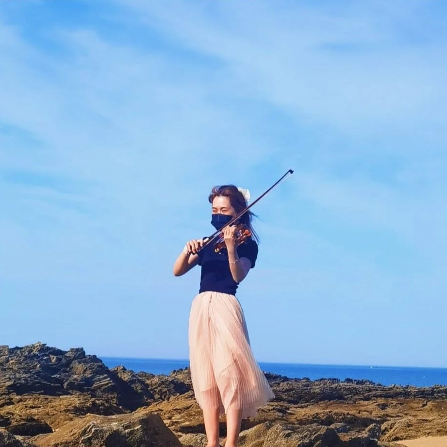 Une femme joue du violon sur la côte rocheuse, portant un masque noir, une jupe rose pâle et un haut noir, avec la mer en arrière-plan.