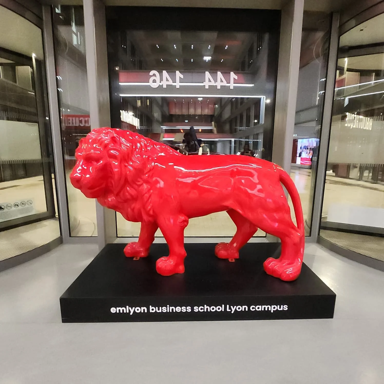 Une sculpture rouge d'un lion en face d'une entrée en verre avec des inscriptions en miroir derrière. La sculpture est placée sur un socle noir avec l'inscription "emlyon business school Lyon campus".