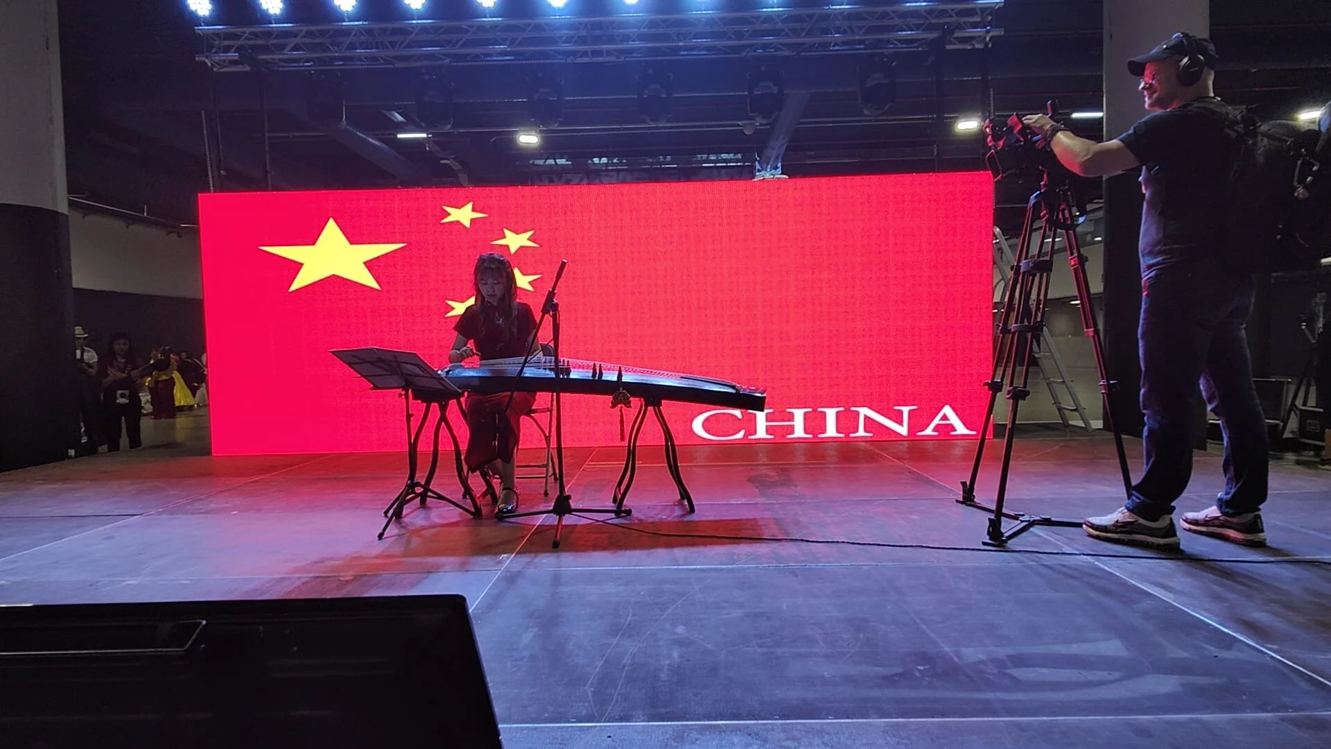 Une femme joue du guqin sur scène, en arrière-plan un écran rouge avec l'emblème de la Chine et le mot 'CHINA' en blanc, un homme avec une caméra enregistre la performance.