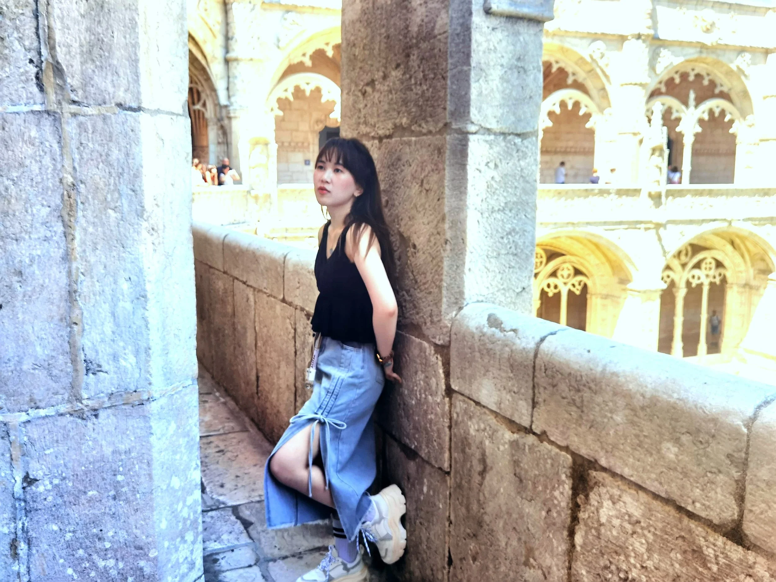 Une jeune femme au cheveux noirs, portant un haut noir avec des bretelles et un pantalon en jean, pose dans un site historique avec des arches en pierre.