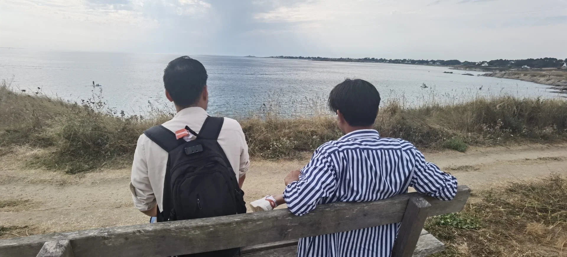 Deux jeunes hommes assis et regardant la mer depuis un rivage rocheux, avec un ciel nuageux et des pêcheurs en bateau au loin.