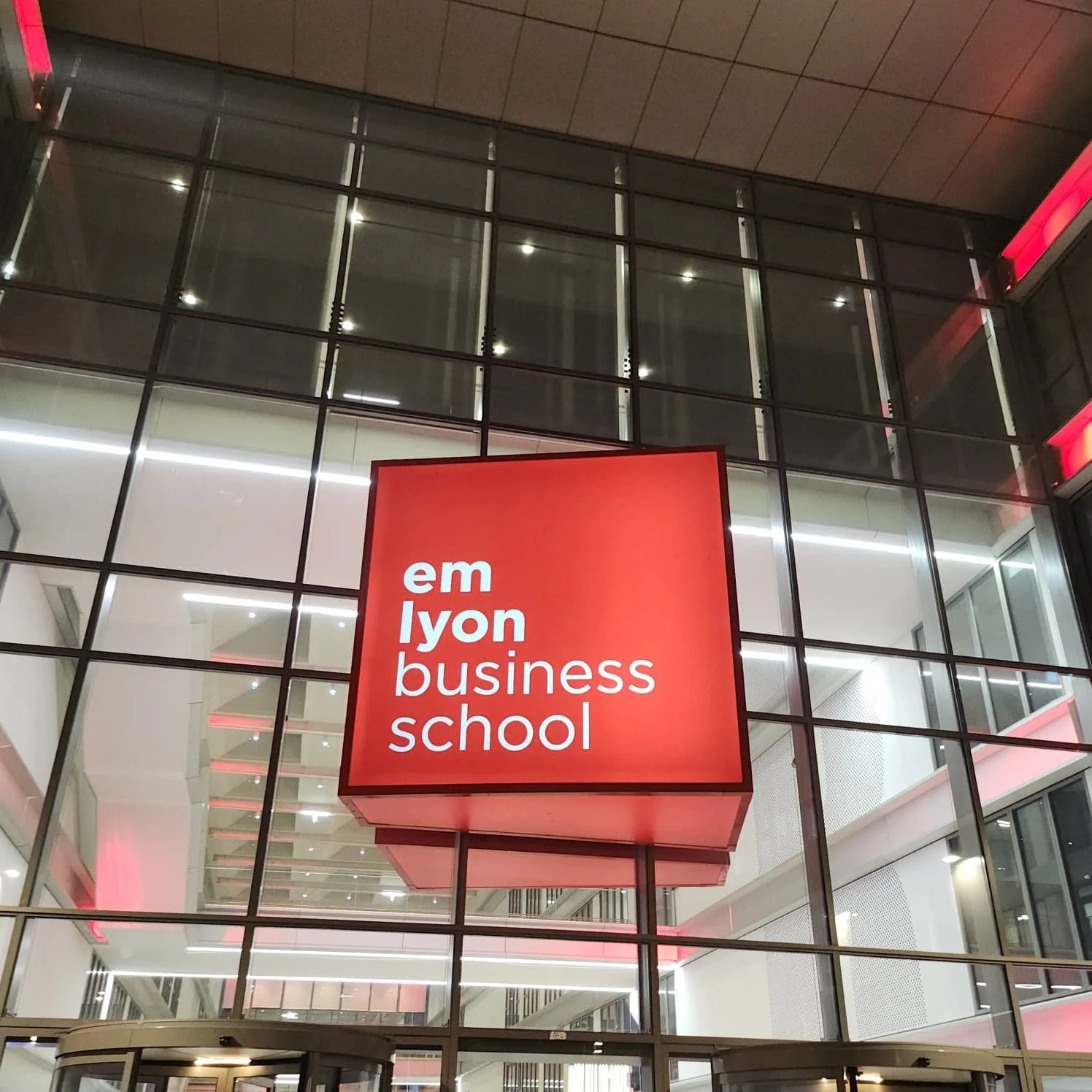 Un bâtiment moderne avec une façade en verre et un panneau rouge indiquant 'emlyon business school'
