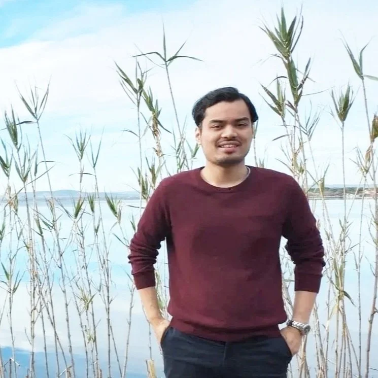Un homme debout devant un lac avec des plantes hautes, portant un pull rouge bordeaux et une montre, avec un ciel nuageux en arrière-plan.