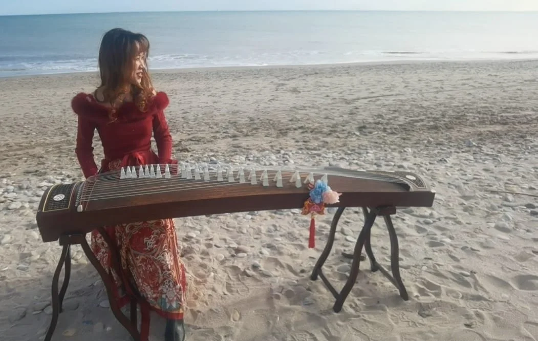 Une femme en vêtements rouges joue du guzheng sur la plage, avec la mer en arrière-plan.