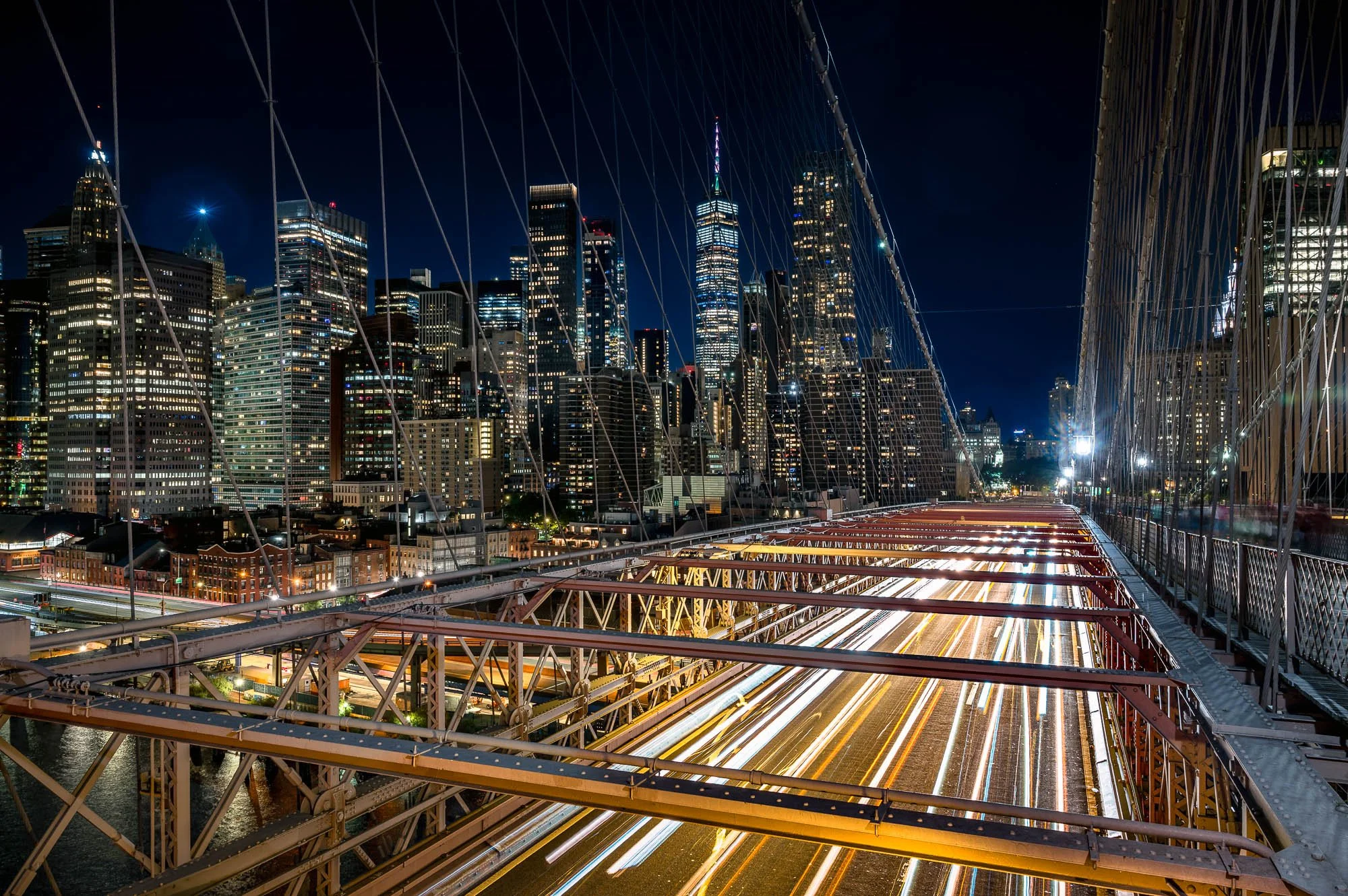 Nighttime view from a bridge overlooking a busy city street with streaks of car lights and illuminated skyscrapers in the background.