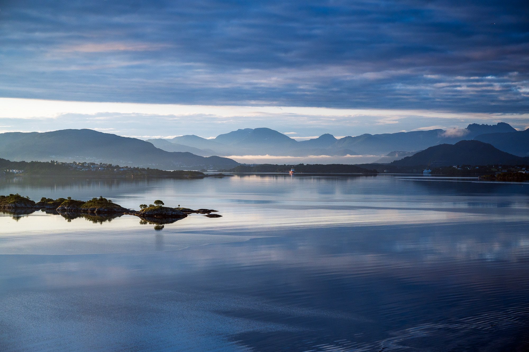 Scenic view of a calm body of water reflecting the cloudy sky above, with distant mountains and small islands along the shoreline.