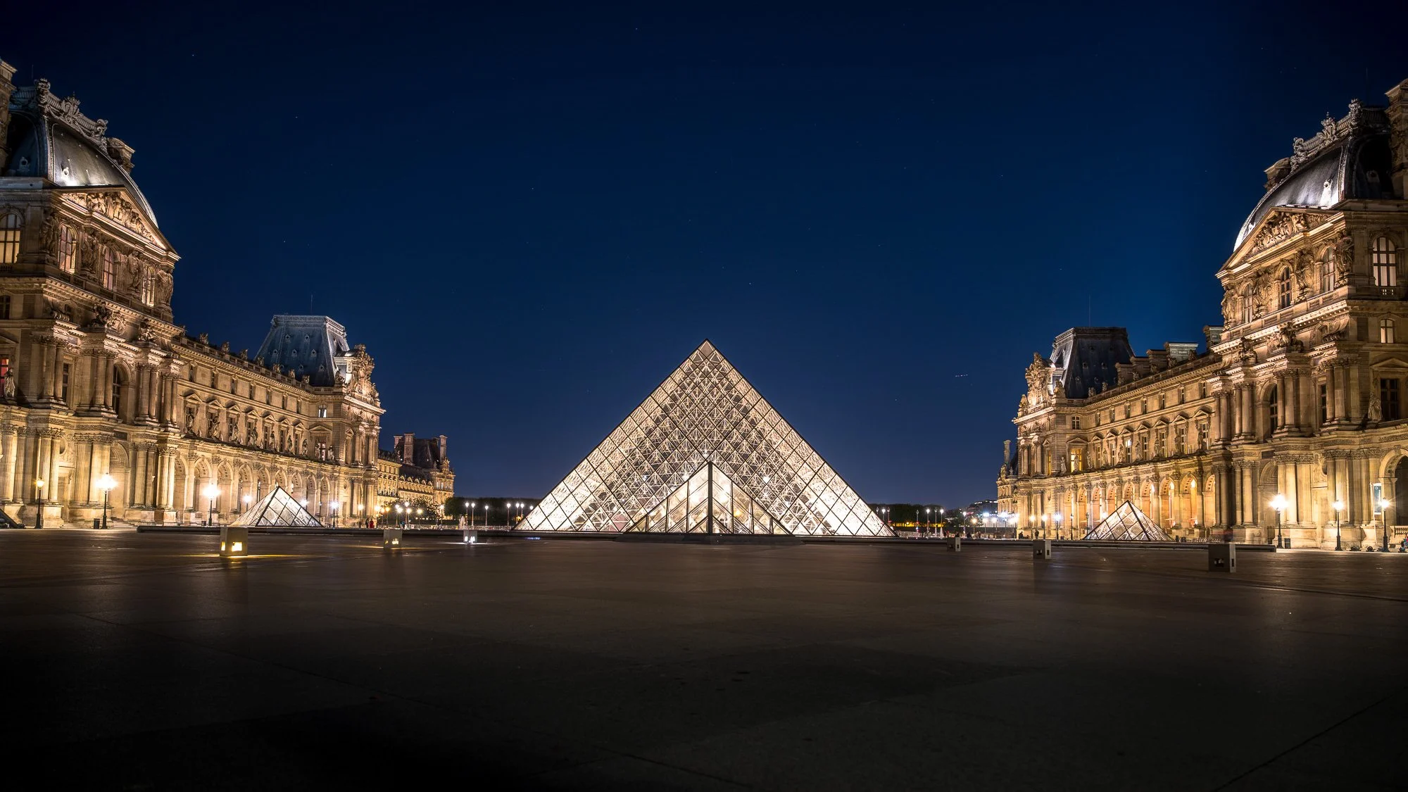 Night view of the Louvre Museum in Paris, France, with illuminated glass pyramid entrance and historic buildings on either side.