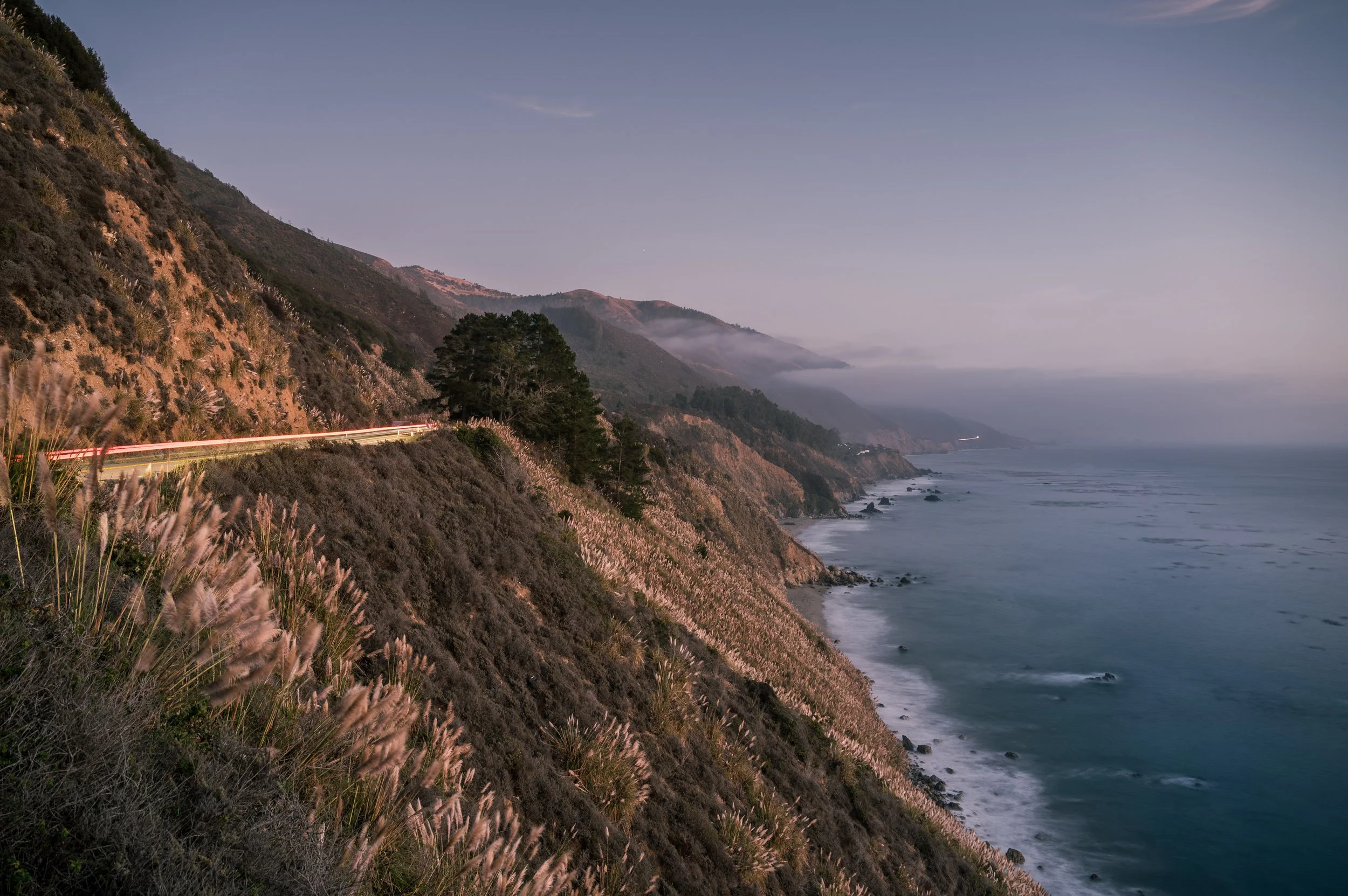 Coastal cliffs with vegetation and a winding road, overlooking the ocean with waves and a cloudy sky.