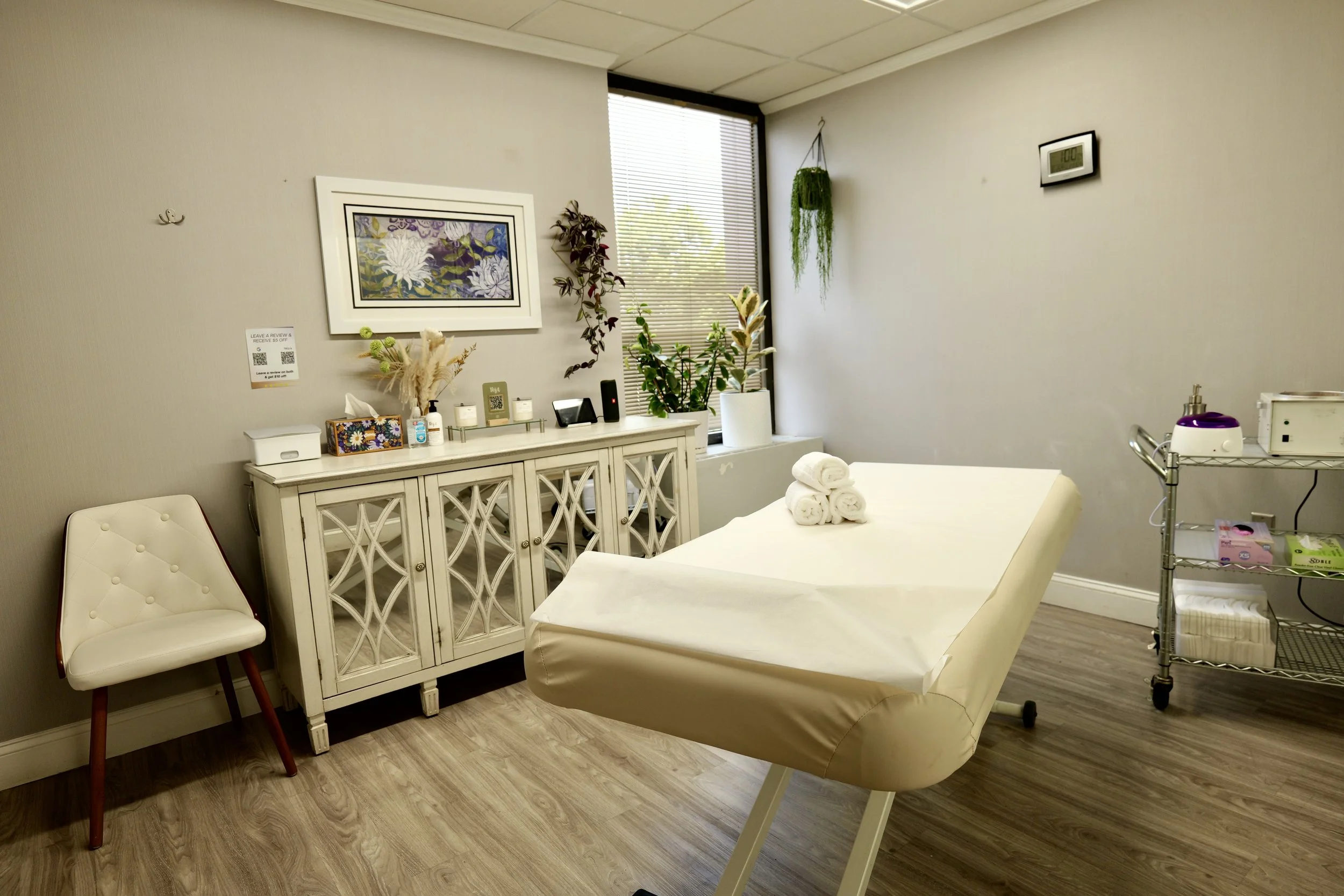 A medical examination room with a padded treatment bed covered with white paper and rolled white towels, a white chair with red legs, a white cabinet with decorative doors, a window with blinds, and various plants and artwork on the walls.