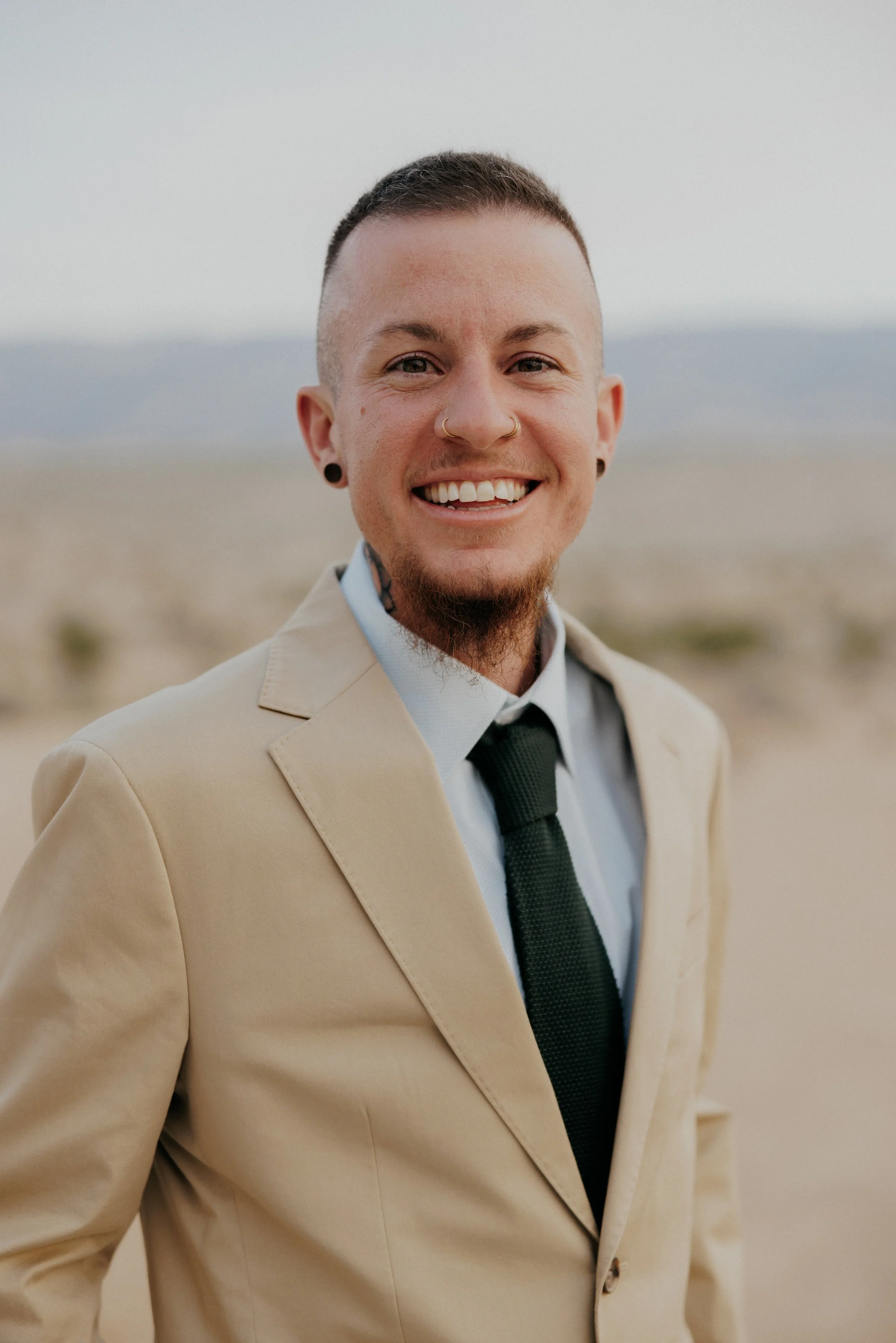 Smiling man in beige suit, white shirt, black tie, standing outdoors in a desert landscape.