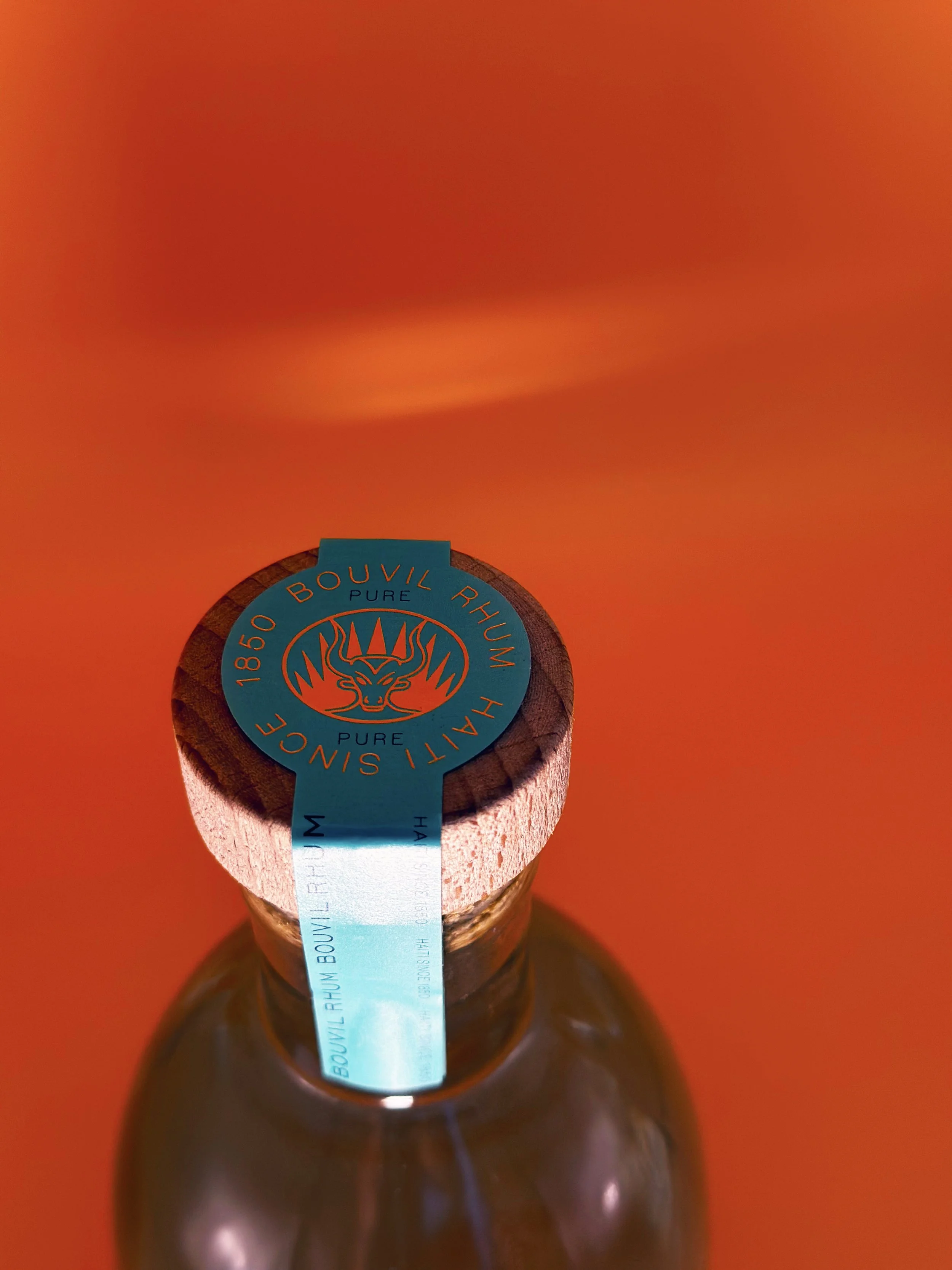 Close-up of a brown glass bottle with a wooden cap, sealed with a blue label that reads "1805 Bouvil Rhum" and features a lion's head logo, against an orange background.