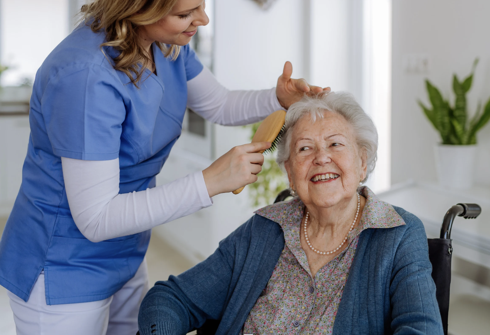 A caregiver brushing the hair of an elderly woman sitting in a wheelchair, smiling, in a well-lit room with houseplants.