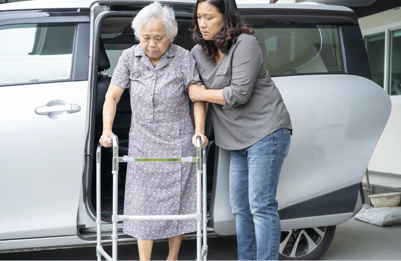 A caregiver assisting an elderly woman with walking using a walker as she exits a vehicle.