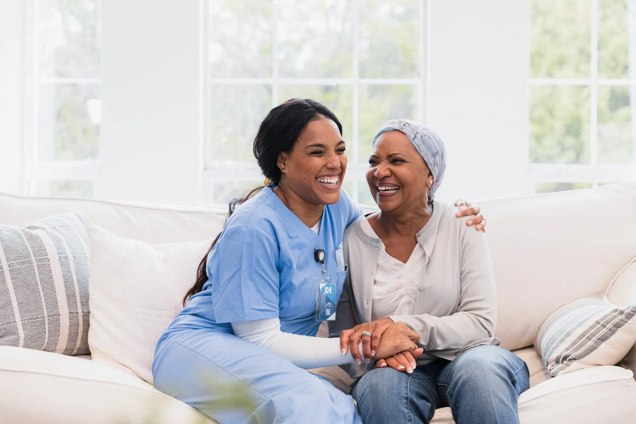A nurse in blue scrubs and a senior woman sit together on a cream-colored couch, smiling and laughing in a bright room with large windows.