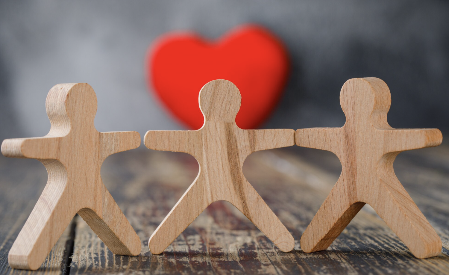 Three wooden human-shaped figurines holding hands with a red heart in the background.