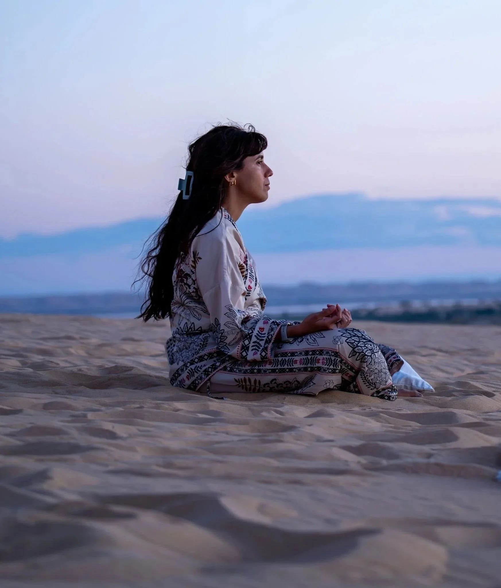A woman sitting cross-legged on a sandy beach with her hands resting on her knees, meditating or practicing mindfulness during twilight with a calm ocean and a distant mountain range in the background.