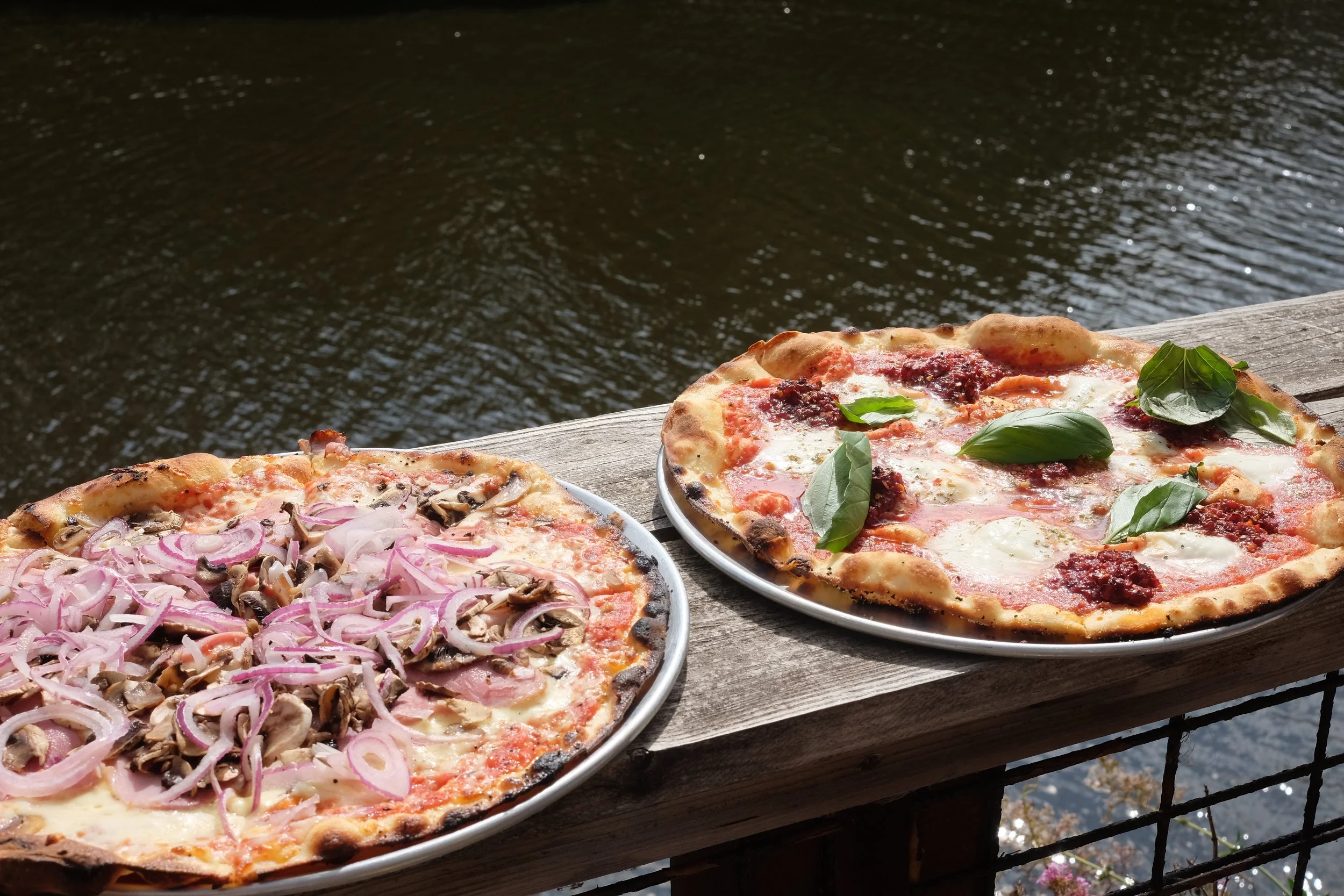 Two delicious pizzas on plates resting on a wooden railing by a body of water, one with mushrooms and onions, the other with basil leaves and tomato sauce.