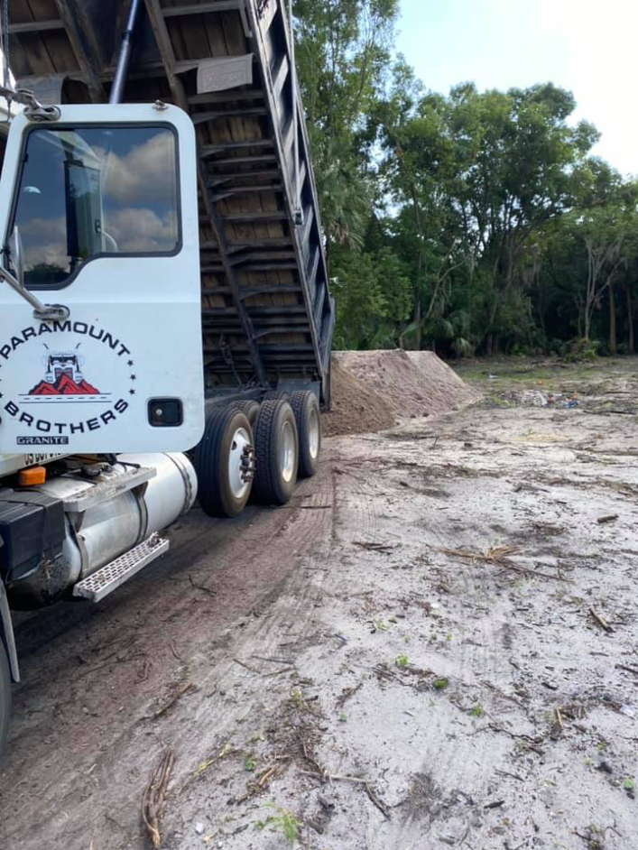 A dump truck with the logo 'Paramount Brothers' on the door, partially raised bed in a construction or dirt removal site, with a dirt pile and trees in the background.