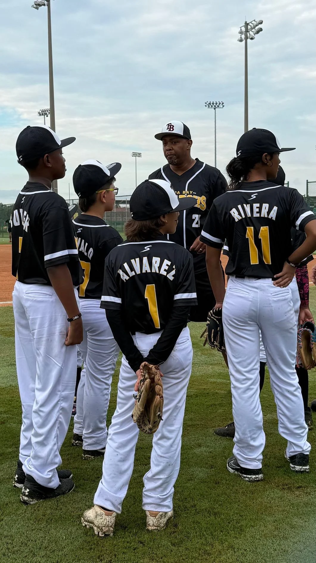 A group of young baseball players wearing black jerseys with white pants, listening to their coach on a baseball field.