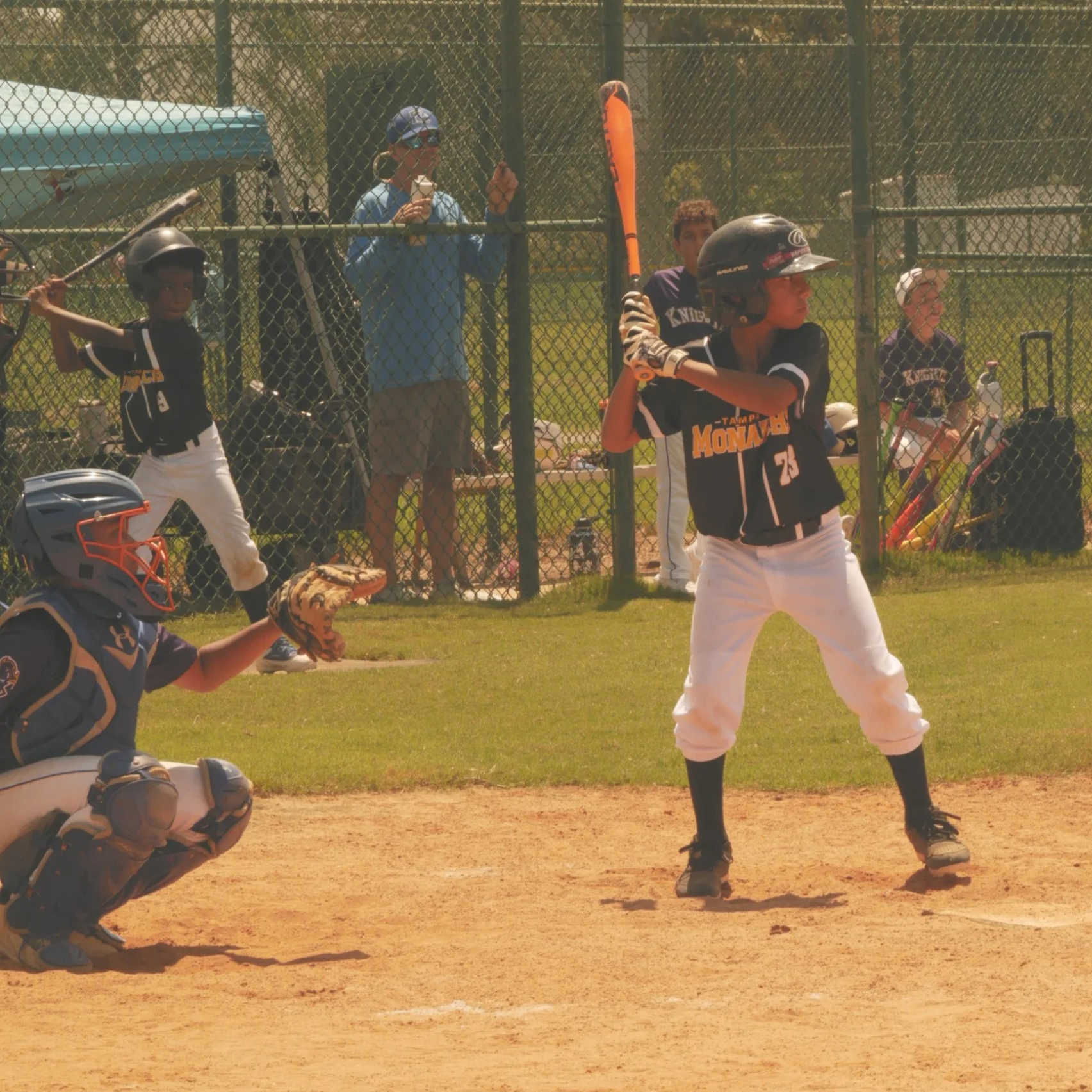 A young baseball player standing at home plate with a bat, ready to swing during a game. The catcher is crouched behind him, and a coach or parent is nearby. Other players and spectators are visible behind the fence.