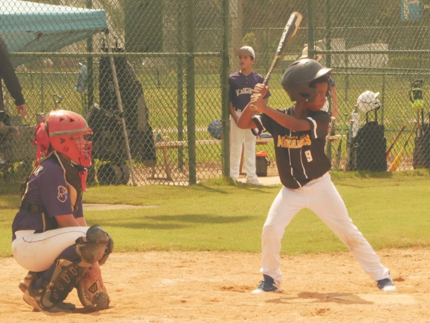 A young baseball player at bat during a game, wearing a helmet and uniform, with a catcher crouching behind him and teammates and coaches watching behind a chain-link fence.