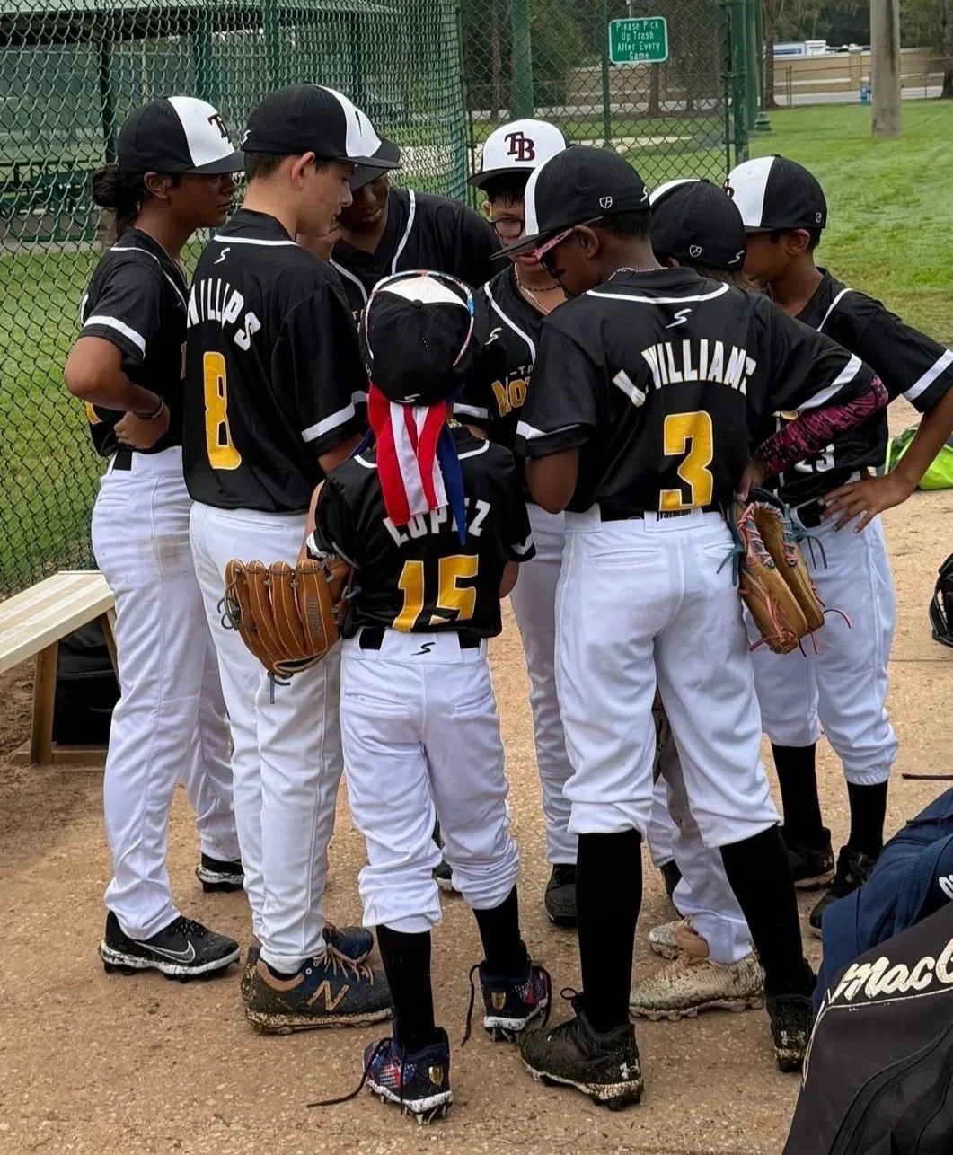 Youth baseball team huddling on the field in black and white uniforms, with a coach or team leader. A sign in the background says, 'Please Pick Up Trash After Every Game.'