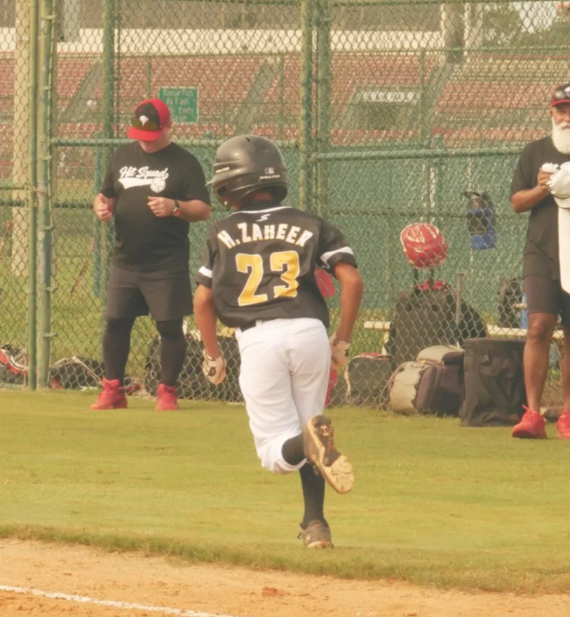 A young baseball player running along the first base line during a game, with coaches or spectators watching from behind a chain-link fence.