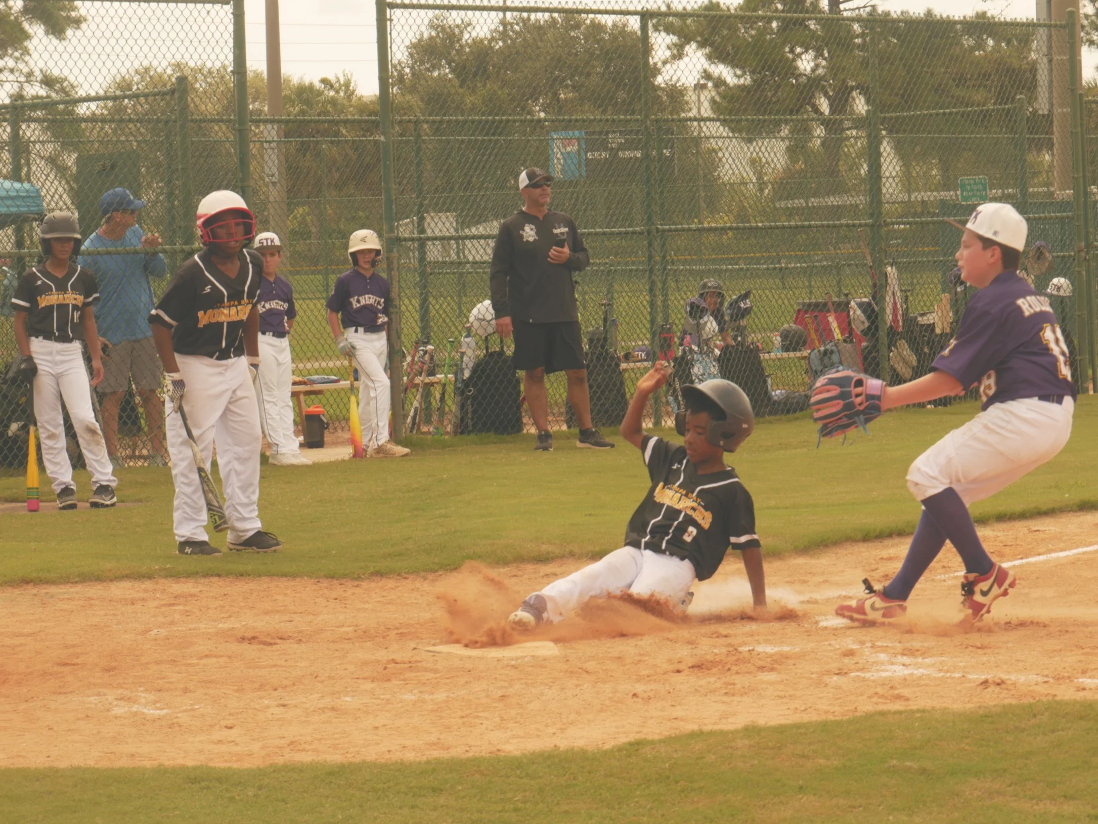 A youth baseball game where a player slides into home plate as a catcher attempts to tag him out. Several other players and coaches watch from the background.