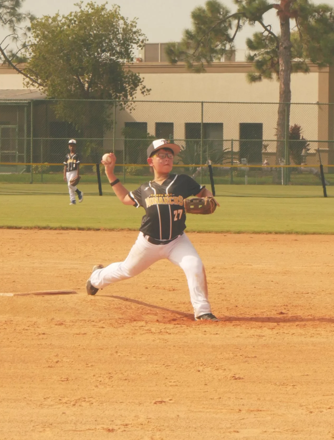 A young baseball player in a black and white uniform is pitching on a baseball field, with a grassy outfield, trees, and a building in the background.