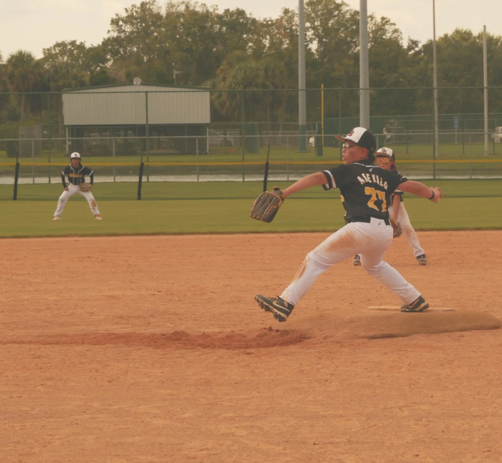 A young baseball player wearing a black and yellow uniform and cap is in the process of catching a ball on a baseball field. Other players are visible in the background, with one near the outfield fence and another behind the player.