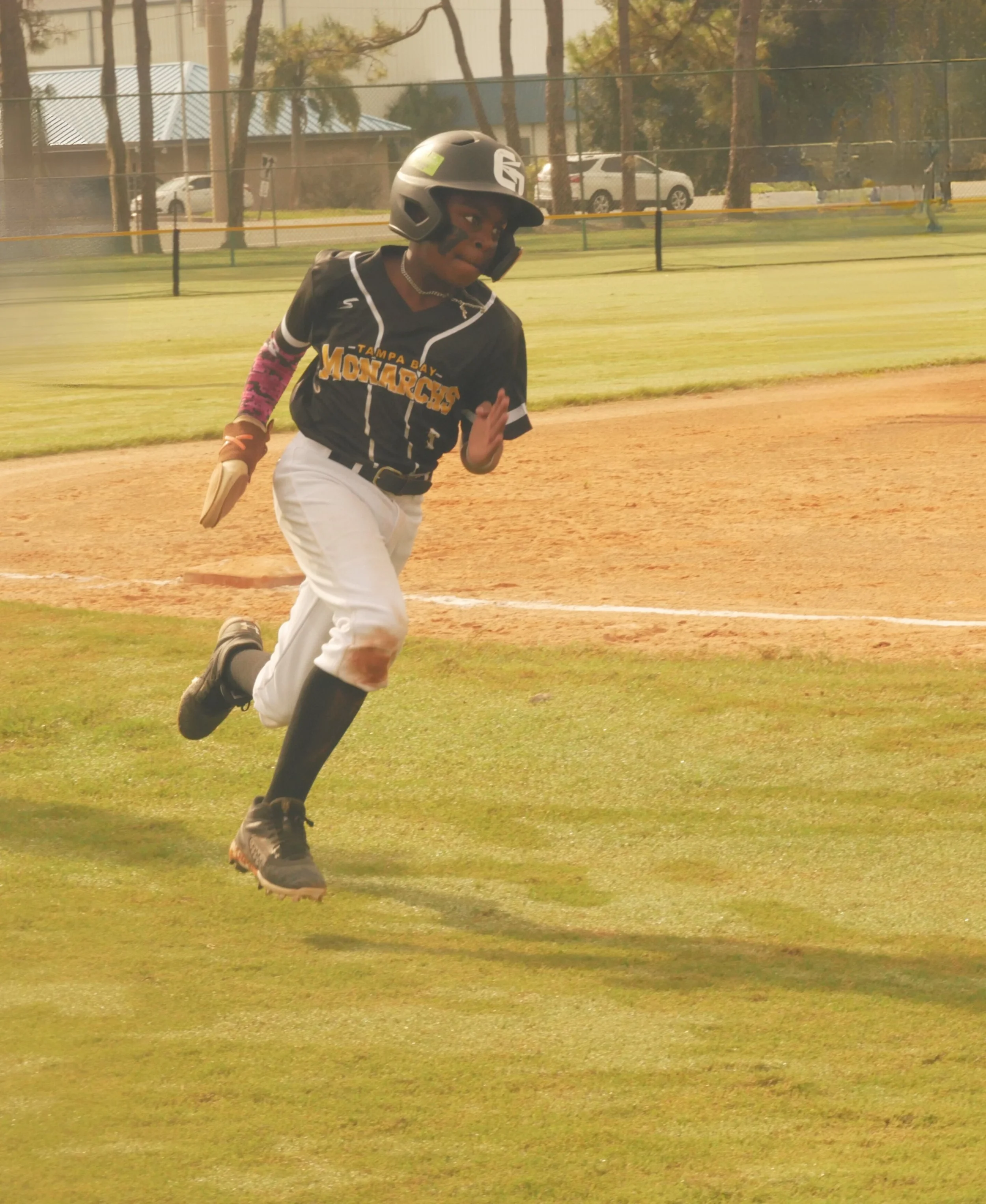 Young baseball player running on the field during a game, wearing a black helmet, black jersey, white pants, and black cleats.