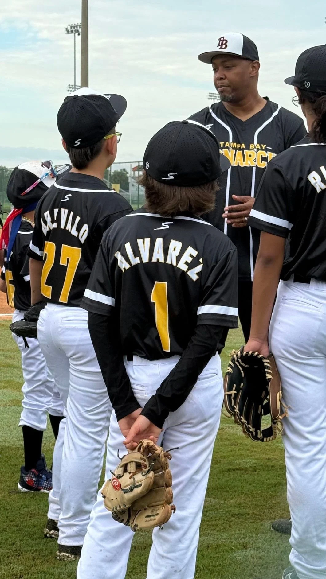 A youth baseball team listening to their coach during a game or practice, wearing black and white uniforms with yellow name and number, with a grassy field and light poles in the background.