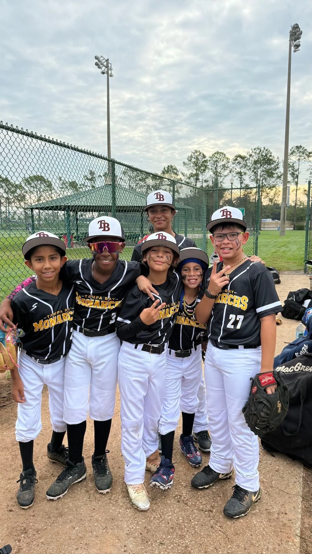 A group of young girls in baseball uniforms and caps, smiling and posing for a photo on a baseball field, with a fence and trees in the background.