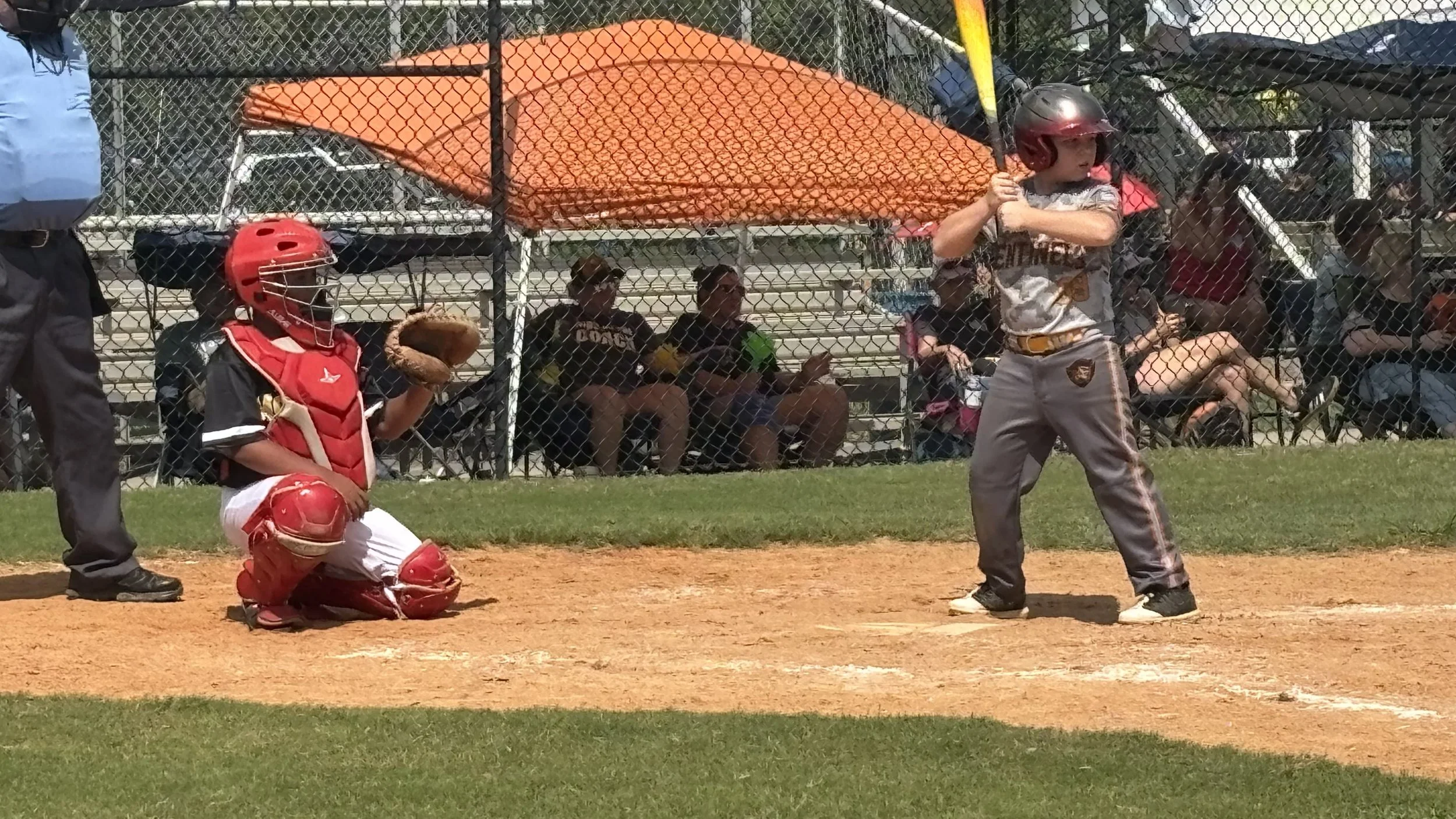 Young baseball player in batting stance at home plate, dressed in a baseball uniform with a gray and gold logo, wearing a helmet, holding a bat, on a baseball field with grass and dirt, with spectators and other players seated behind a chain-link fen