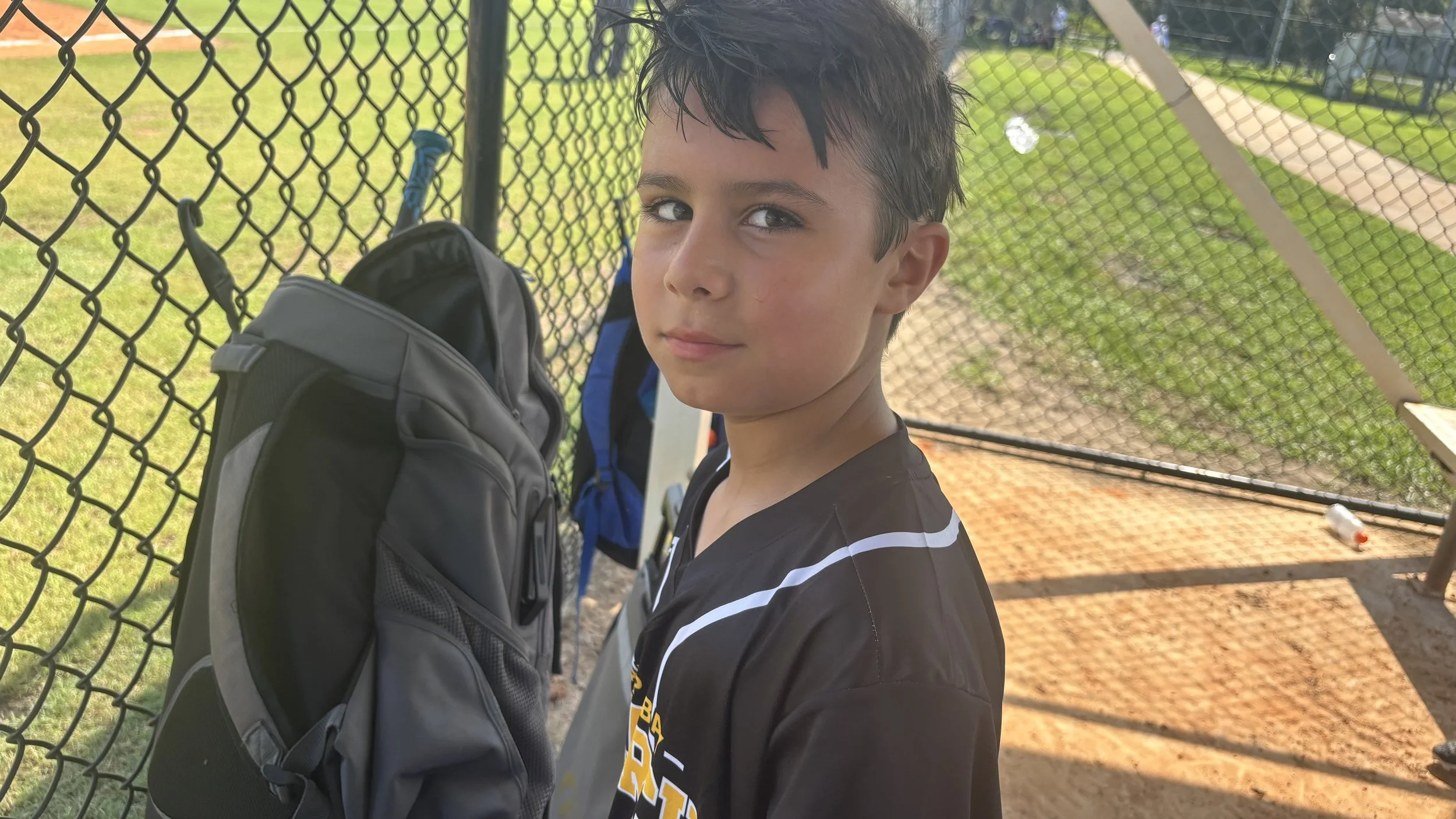 A young boy standing next to a chain-link fence with a baseball backpack, looking at the camera with a serious expression.