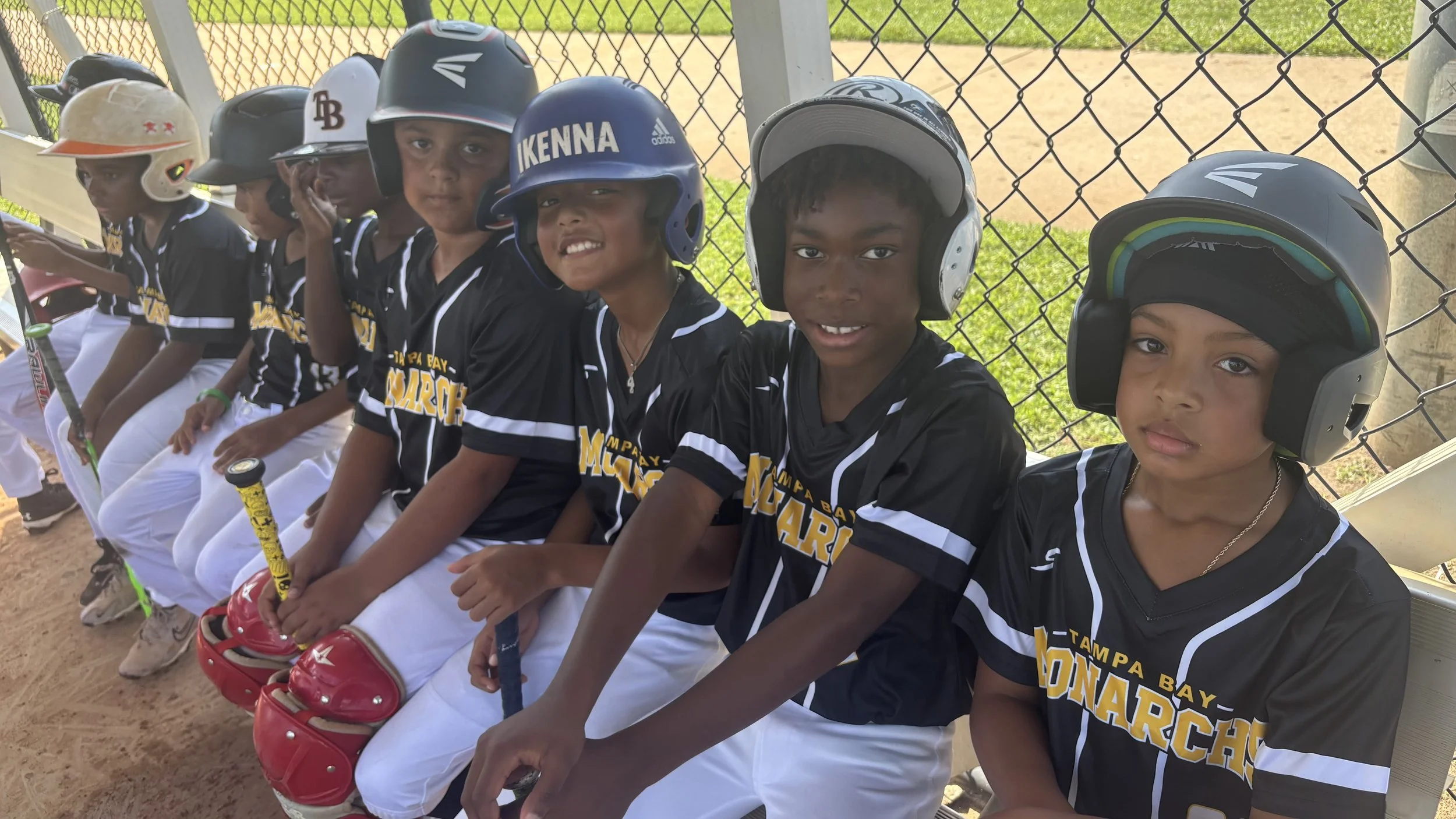 Kids in black baseball uniforms sitting on a bench at a baseball field, wearing helmets, holding bats, with a chain-link fence in the background