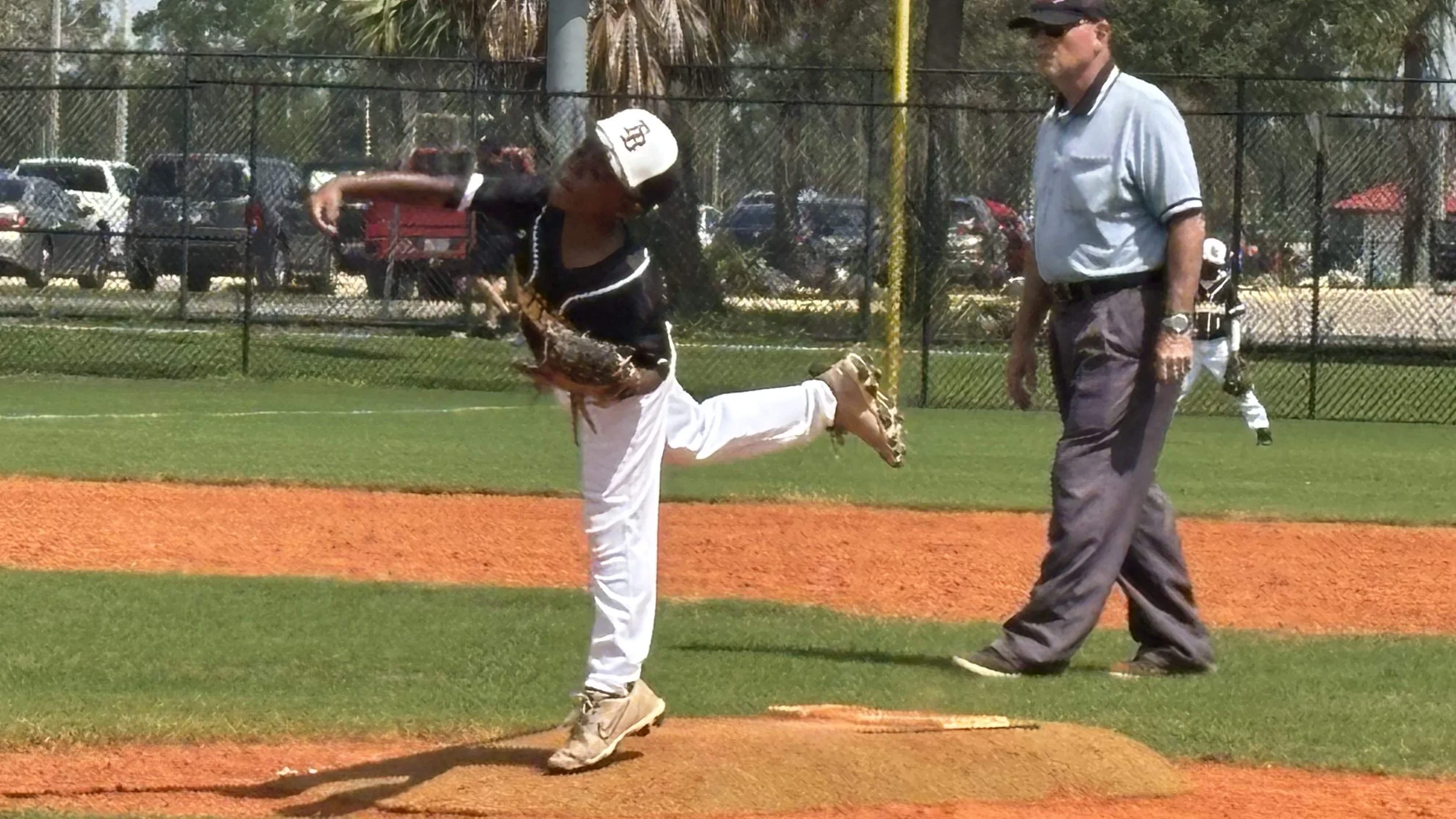A young baseball player is pitching from the mound during a game, with an umpire standing nearby, on a baseball field with a fence and parking lot in the background.