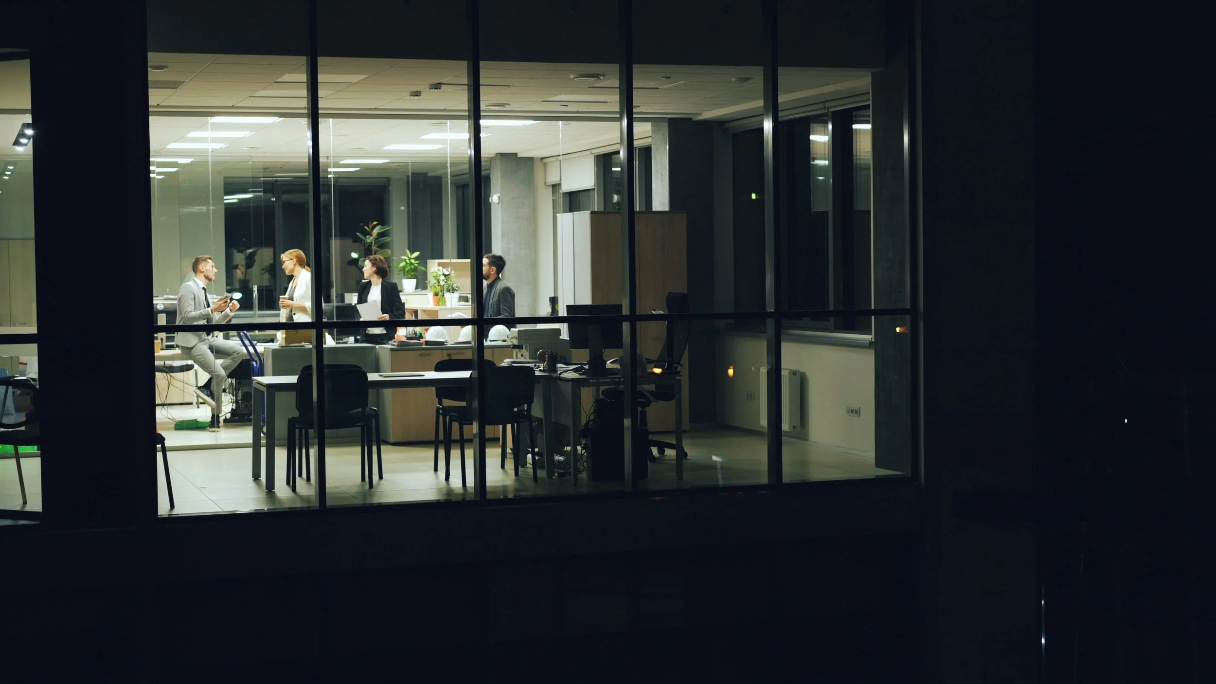 Night view of a modern office with large glass windows showing four people having a discussion inside, with desks and office chairs visible.