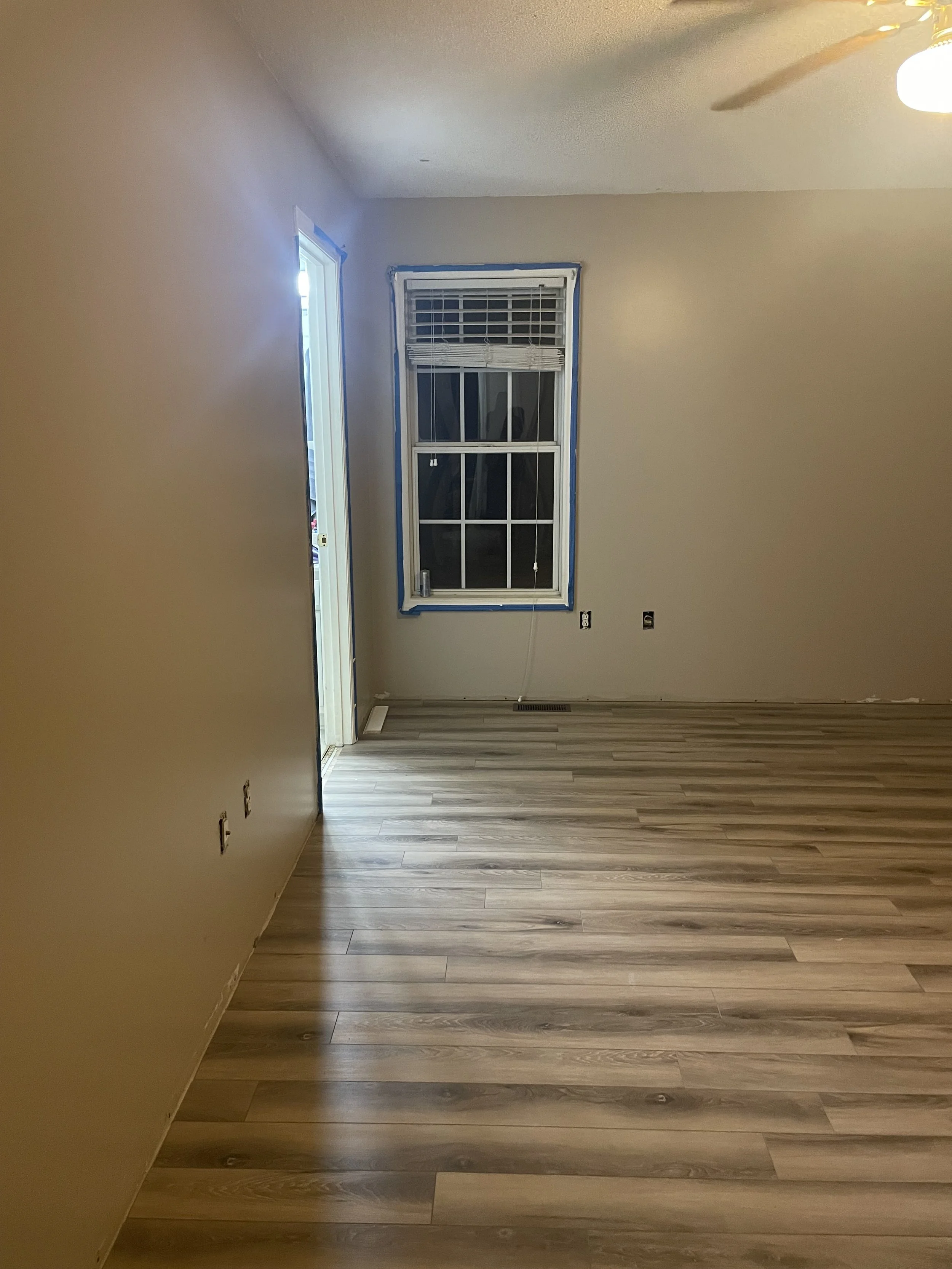 Empty room with beige walls, a window with blinds, and wood laminate flooring. A ceiling fan is partially visible. A door frame leading into another room or hallway shows removal of trim or painting tape around the window.
