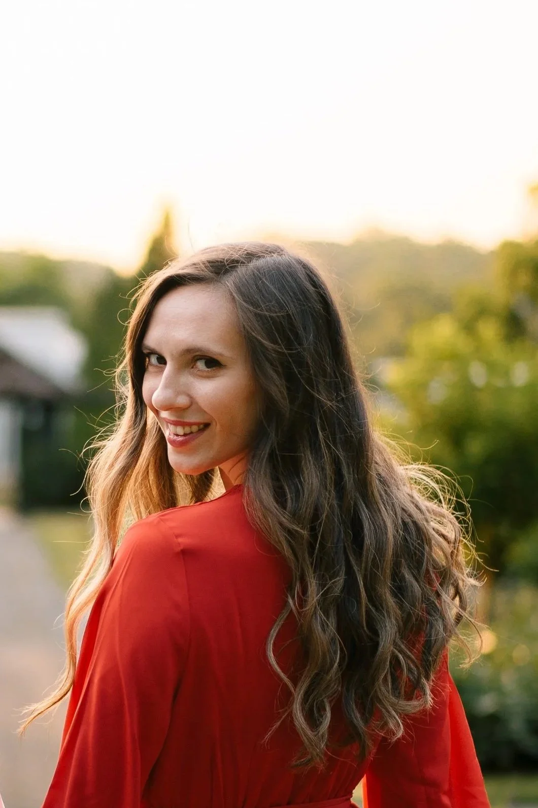 A woman with long, wavy brown hair wearing a red dress looking over her shoulder and smiling outdoors during sunset.