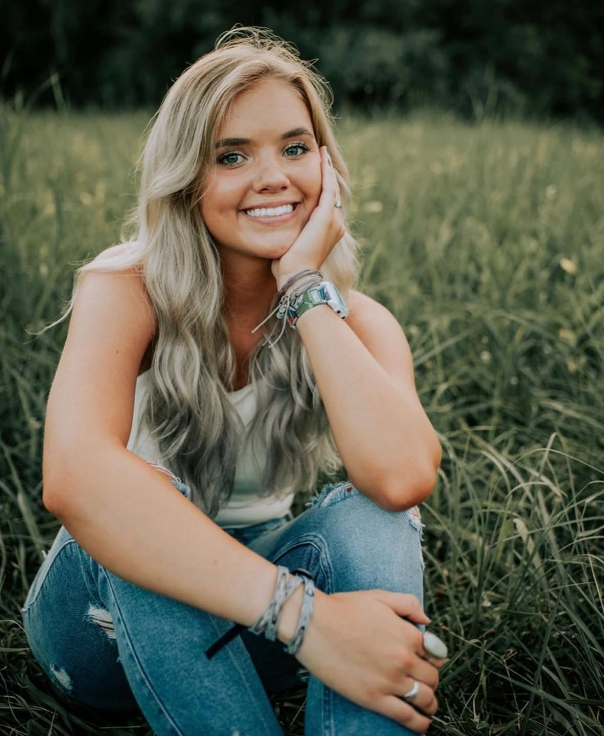 A young woman with long wavy blonde hair, smiling, sitting in a grassy field with trees in the background, wearing a white top and blue jeans.