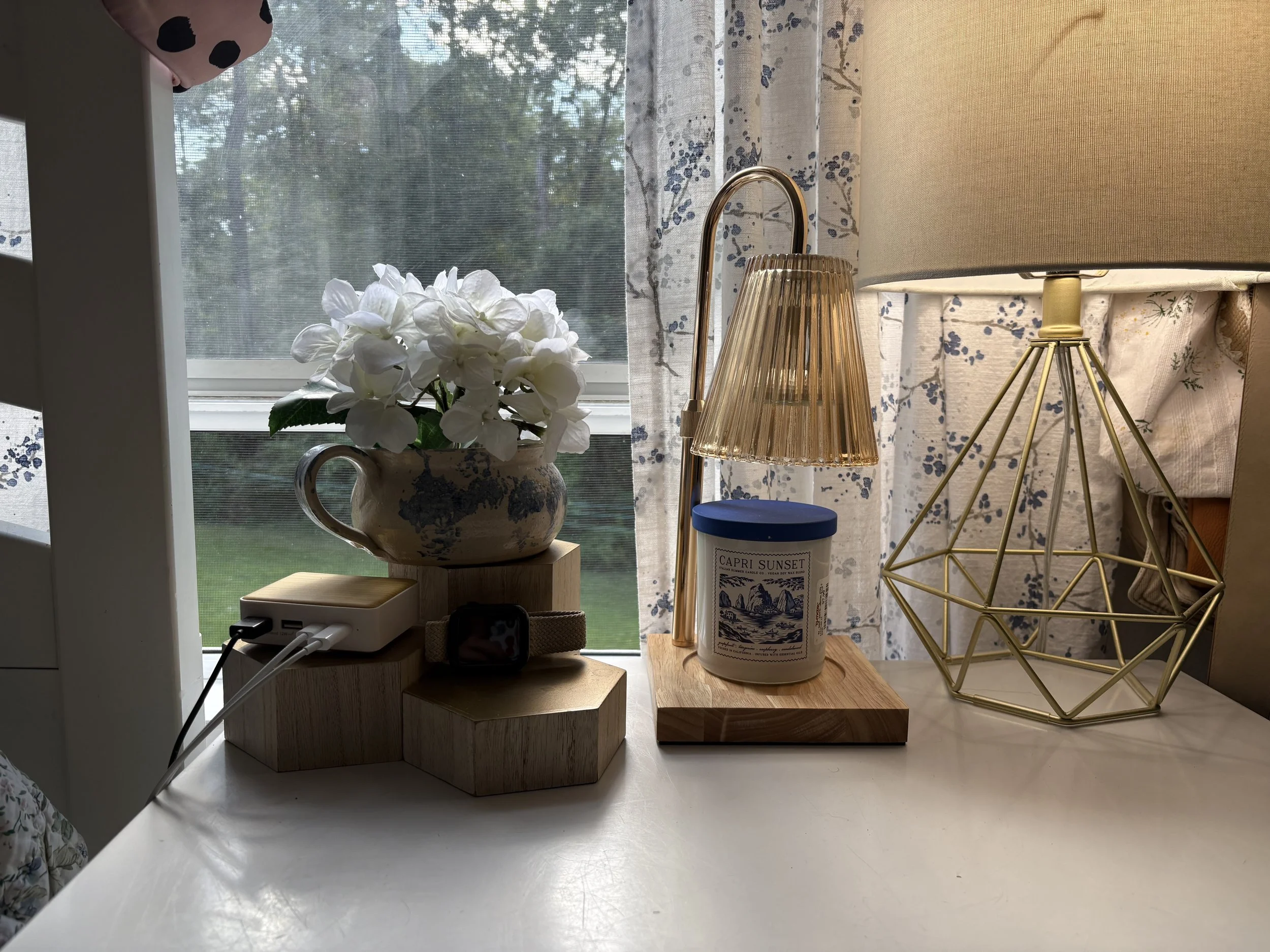 A white table with a wooden block holding a potted white flower, a blue and white scented candle labeled 'Capri Sunset,' a gold geometric table lamp, and a beige lampshade. Behind the items, a window with white and blue floral curtains and a view of greenery outside.
