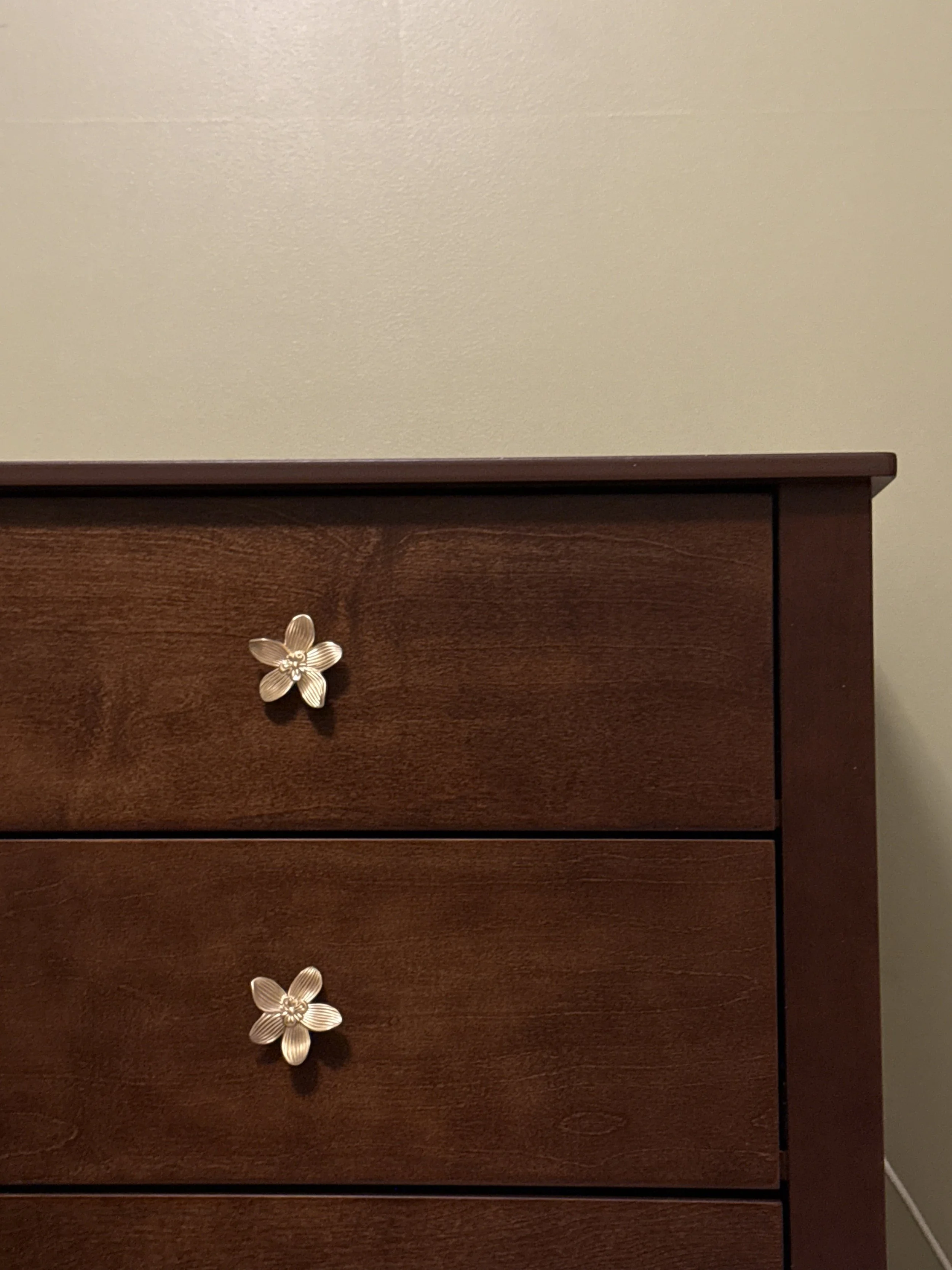 A wooden dresser with three drawers, each with a flower-shaped handle, against a plain beige wall.
