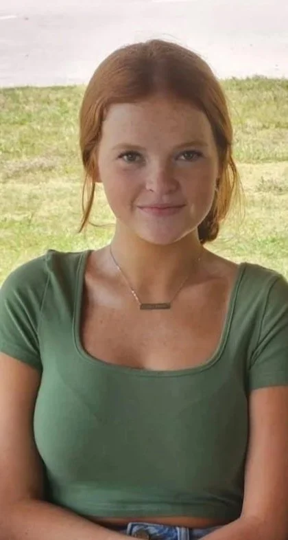 A woman with red hair and a green shirt smiling outdoors with grass and sky in the background.