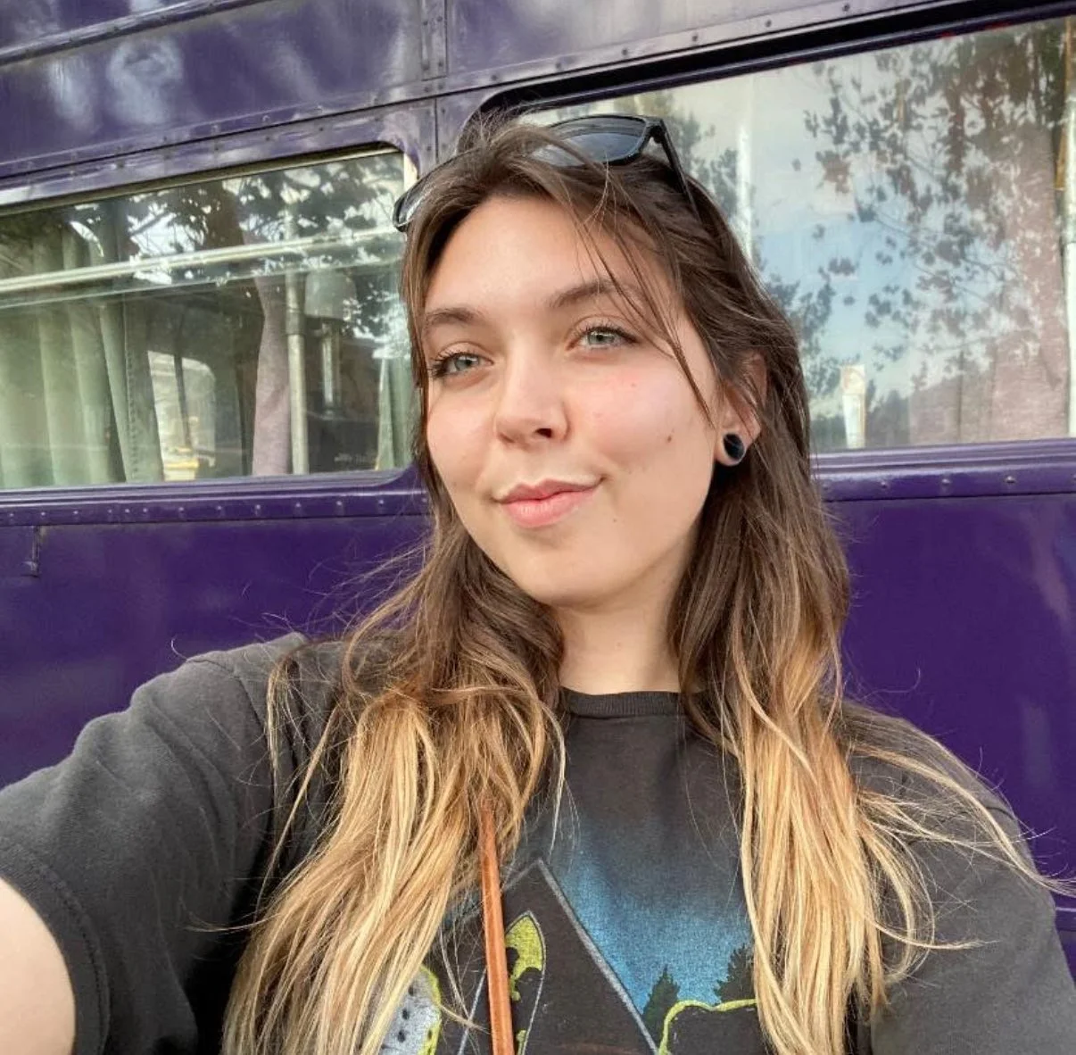 A young woman with long wavy hair, wearing a black T-shirt, sunglasses on her head, and black earrings, taking a selfie in front of a purple bus.