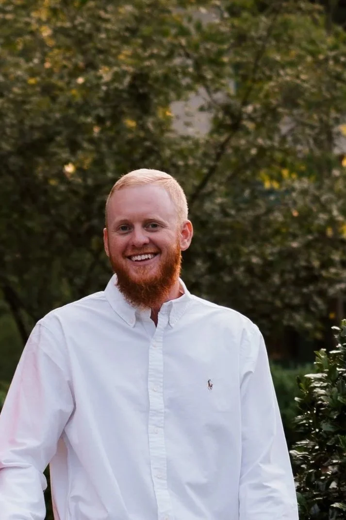 A man with red hair and beard smiling outdoors, wearing a white button-up shirt, with trees in the background.