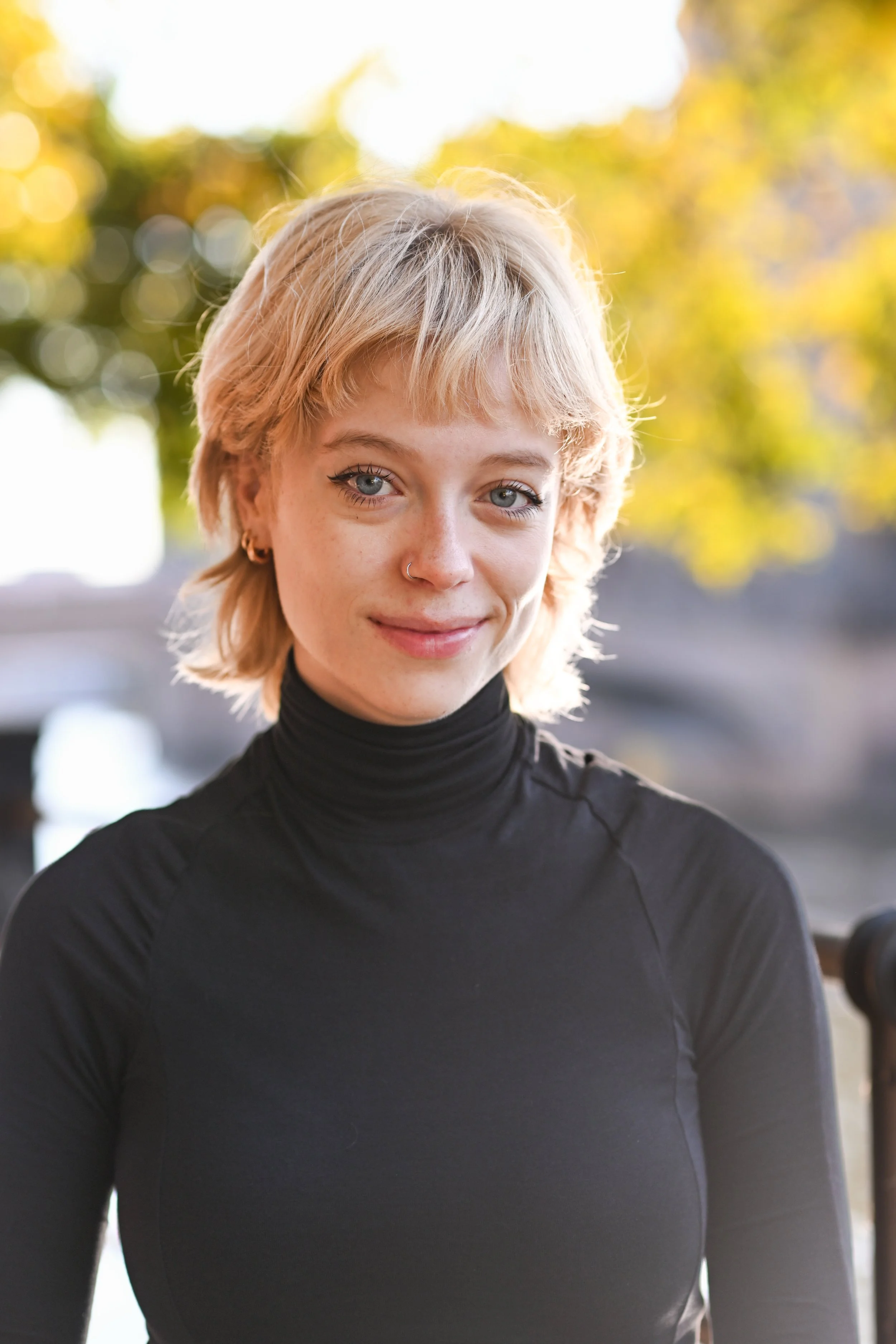A young woman with short blonde hair and blue eyes, smiling, wearing a black turtleneck sweater outdoors during daytime with blurred autumn leaves in the background.